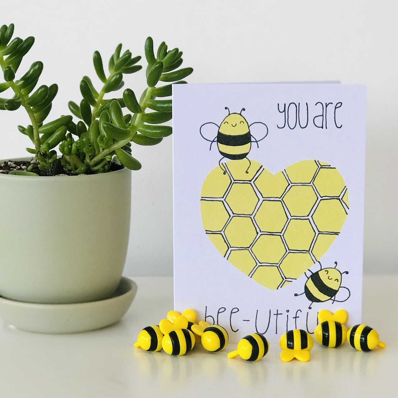 A You Are Bee-utiful Card from Sweet Pea Creations, boasting a hand made print with the pun "you are bee-utiful," stands next to a potted green succulent, accompanied by small bee figurines arranged playfully.