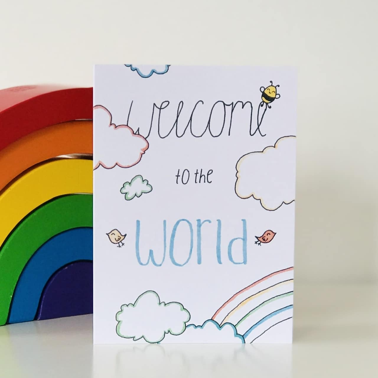 A Welcome to the World Card by Sweet Pea Creations is presented in front of a colorful wooden rainbow toy. The card features playful illustrations of clouds, a sun, and.