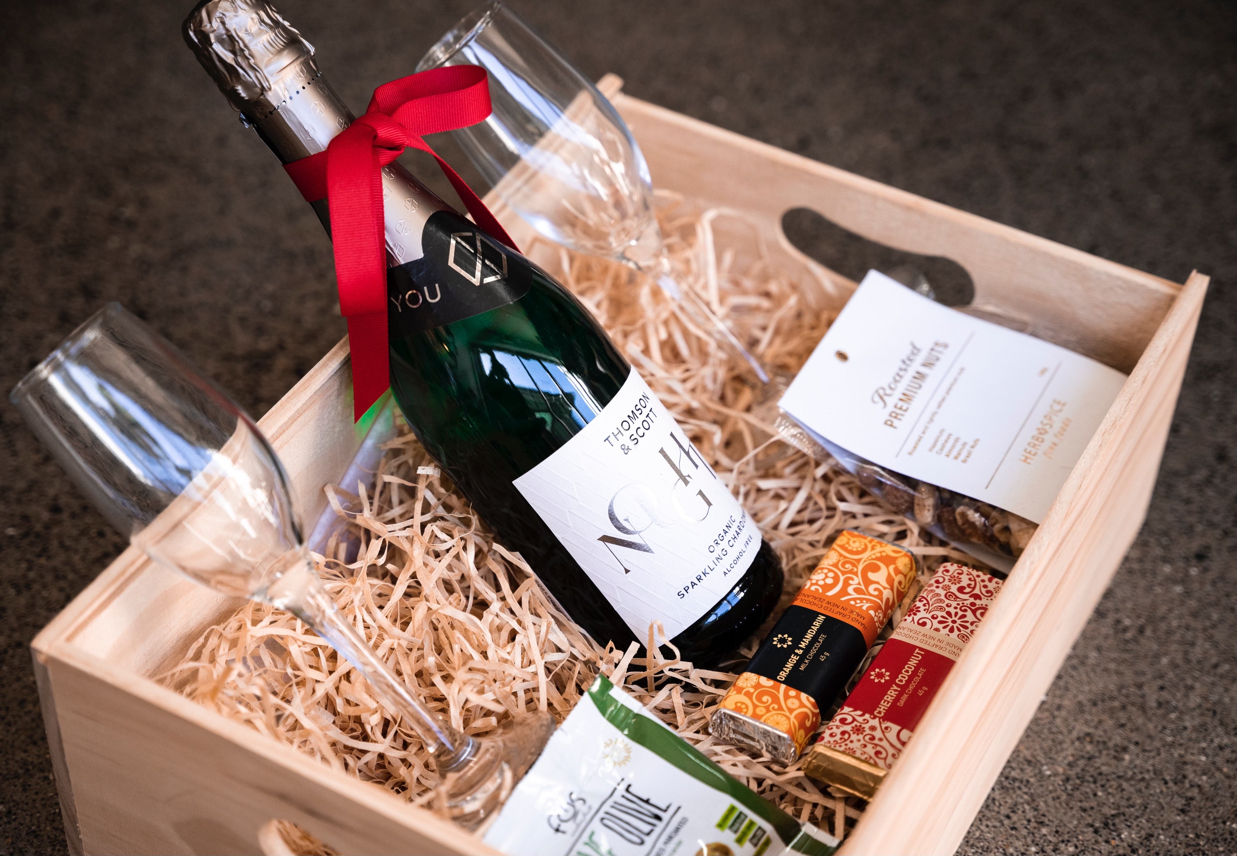 A luxurious Celebrate gift box from giftbox co. with a bottle of non-alcoholic sparkling wine adorned with a red ribbon, two elegant flutes, and an assortment of gourmet snacks carefully presented on a bed of straw.