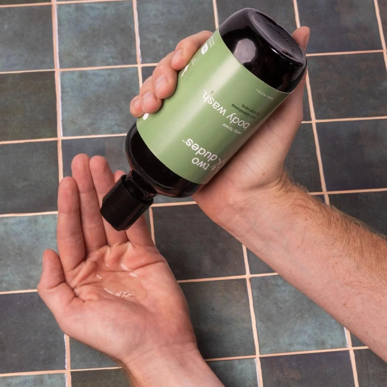 Dispensing Two Dudes kawakawa aloe vera zesty forest body wash into palm over tiled background.