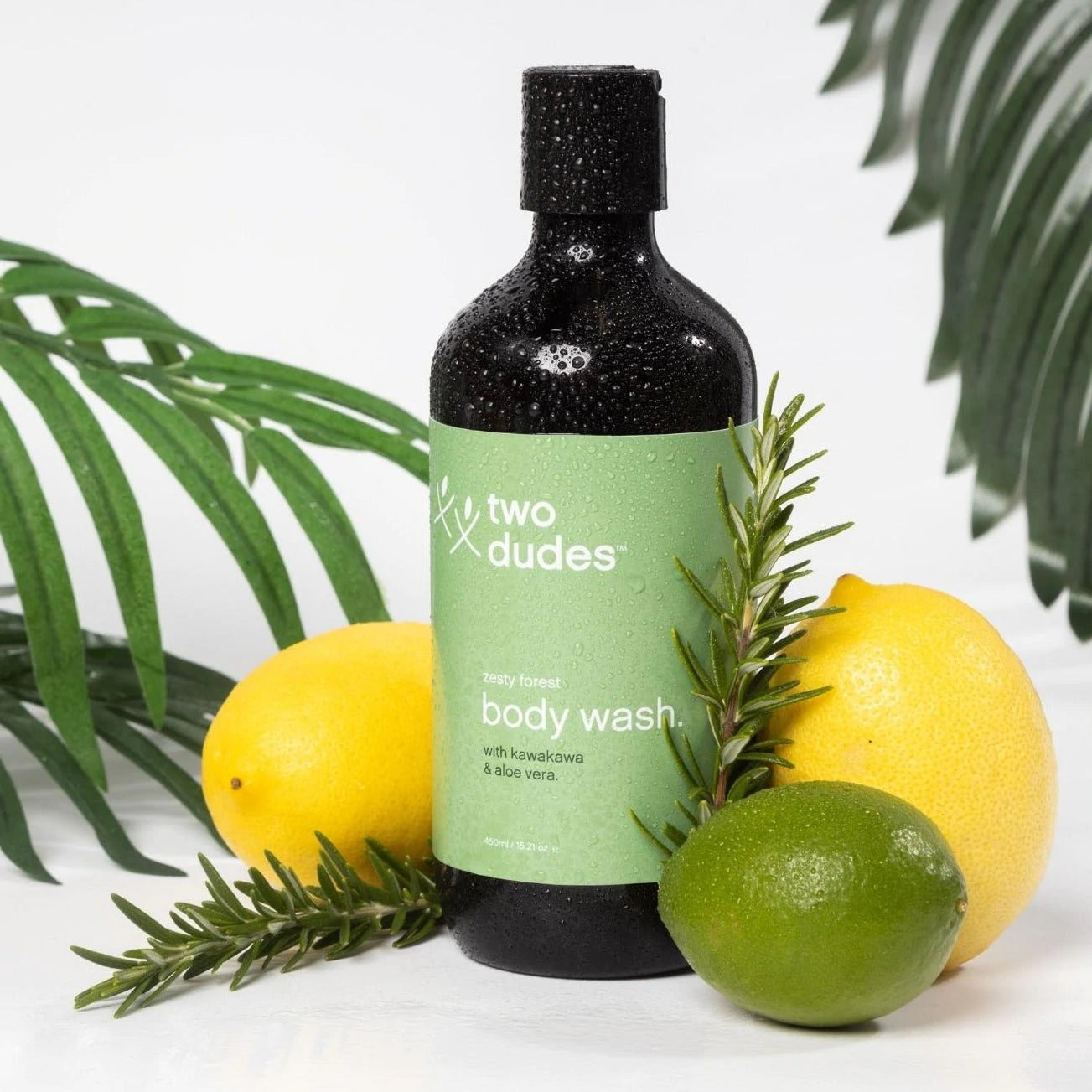 A bottle of "giftbox co. His body wash" with a green label, surrounded by fresh lemons, a lime, and sprigs of rosemary against a white background.