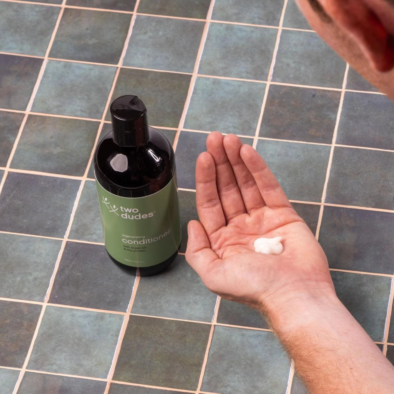 Dispensing a dollop of Two Dudes argan oil regenerating conditioner from a pump bottle onto an open palm over a tiled surface.