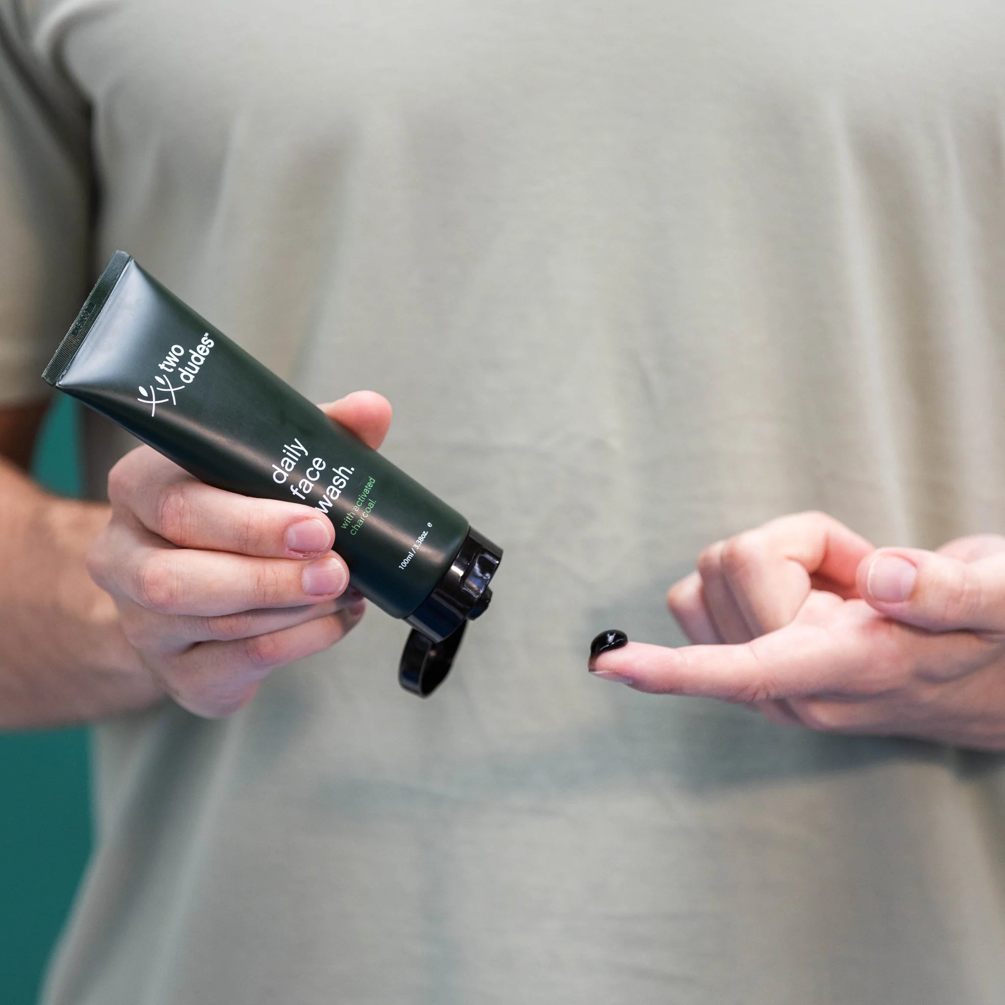 A person wearing a gray t-shirt holds a green tube of Two Dudes Daily Face Wash, squeezing a small amount onto their fingertip.