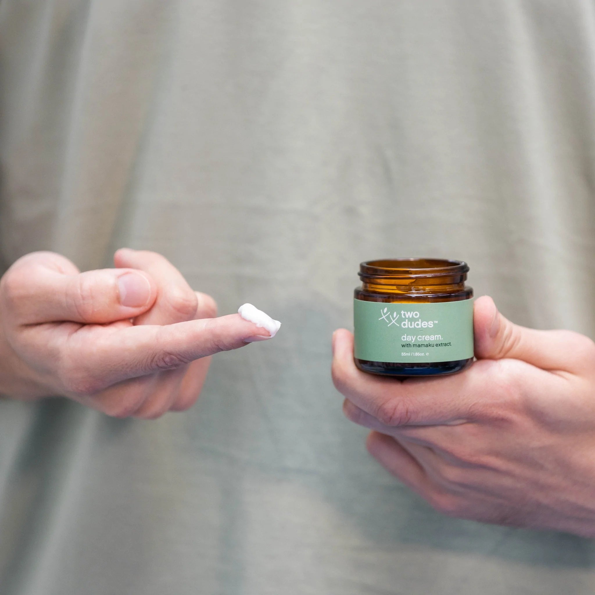 A person holding a jar of "Day Cream by Two Dudes" skin protection day cream in one hand with a dab of the cream on the index finger of the other hand, ready for application.