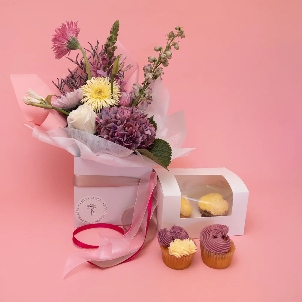 A bouquet of seasonal blooms wrapped in white paper set against a pink backdrop, accompanied by a Frida + Sweet gluten-free floral gift box of cupcakes with yellow frosting.