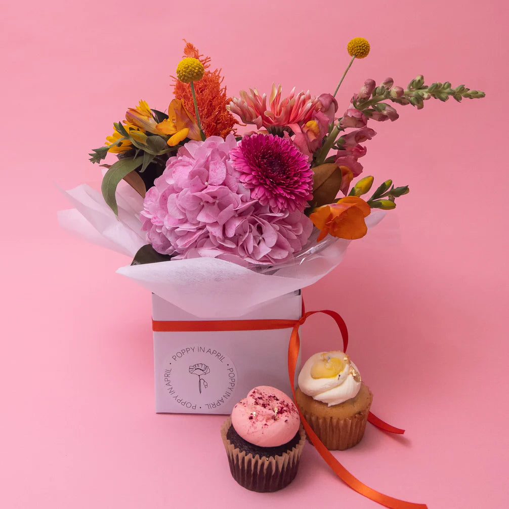 A vibrant bouquet of seasonal blooms in varying shades of pink, orange, and yellow, alongside two Frankie + Sweet cupcakes, one with pink frosting and the other with a swirl of yellow, presented tastefully against Poppy in April.