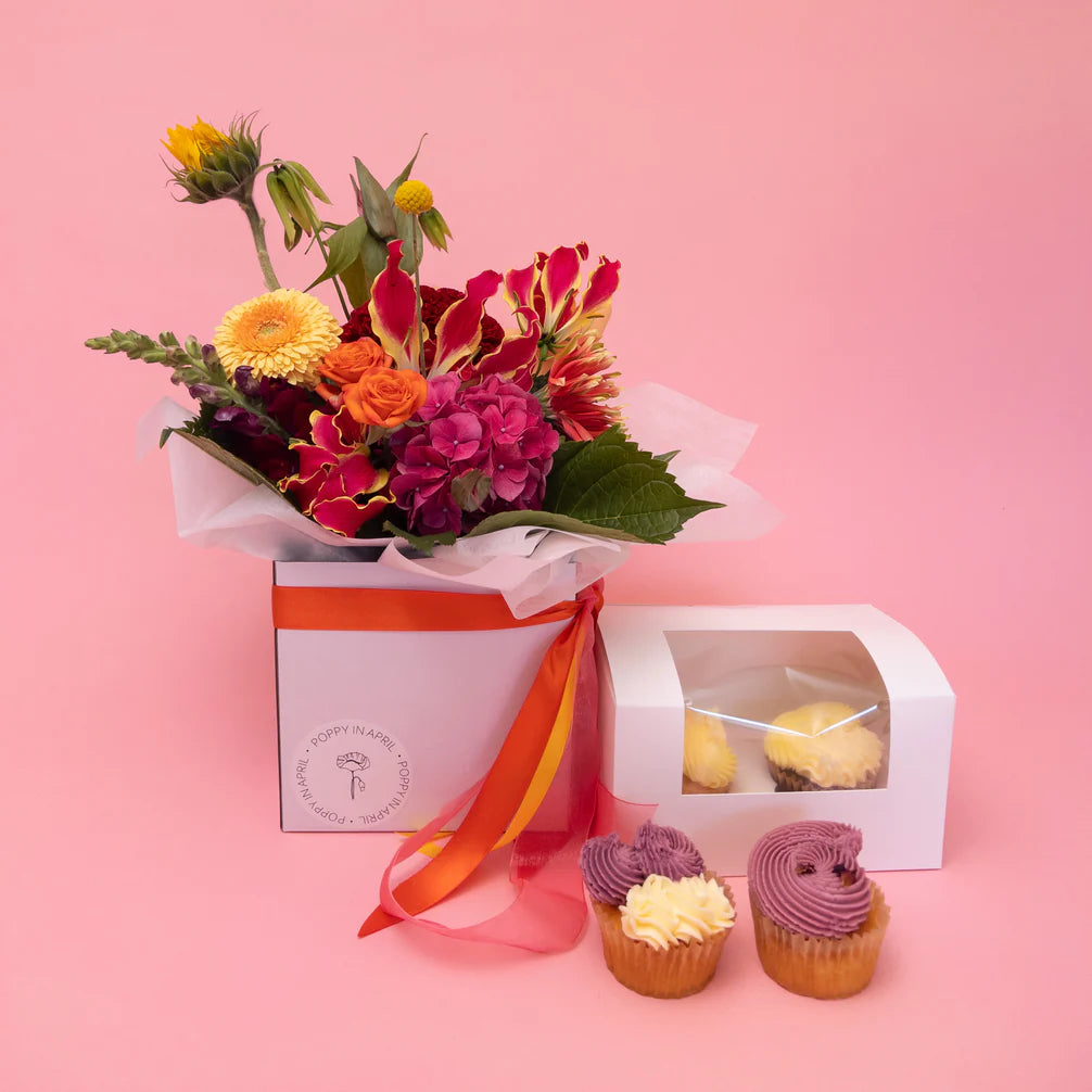 A vibrant bouquet of Frida + Sweet seasonal blooms paired with a box of delightful Poppy in April gluten-free cupcakes against a pink background, offering a feast for the senses.