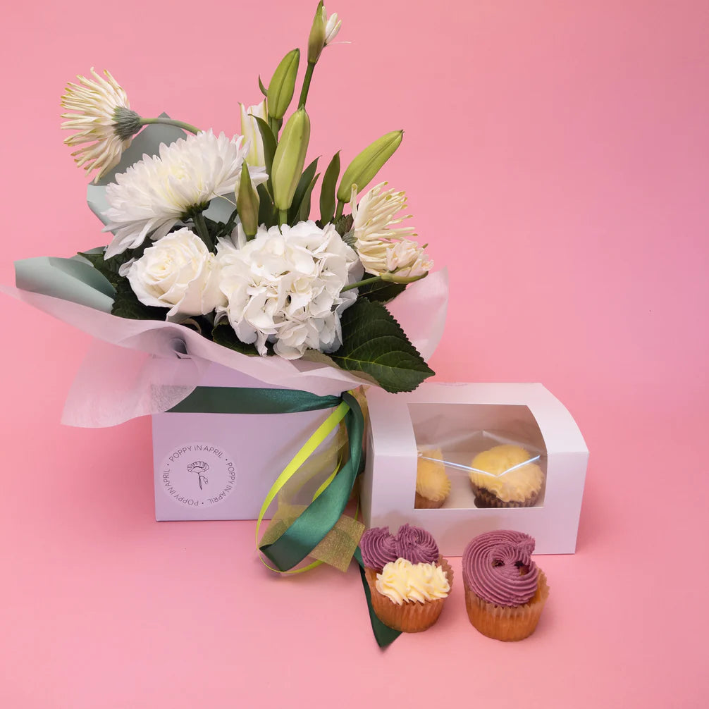 A beautiful bouquet of seasonal blooms with green ribbon next to a Frida + Sweet floral gift box containing two gluten-free cupcakes, and two additional frosted gluten-free cupcakes displayed in front of it, all against a soft pink backdrop. Brand Name: Poppy in April.