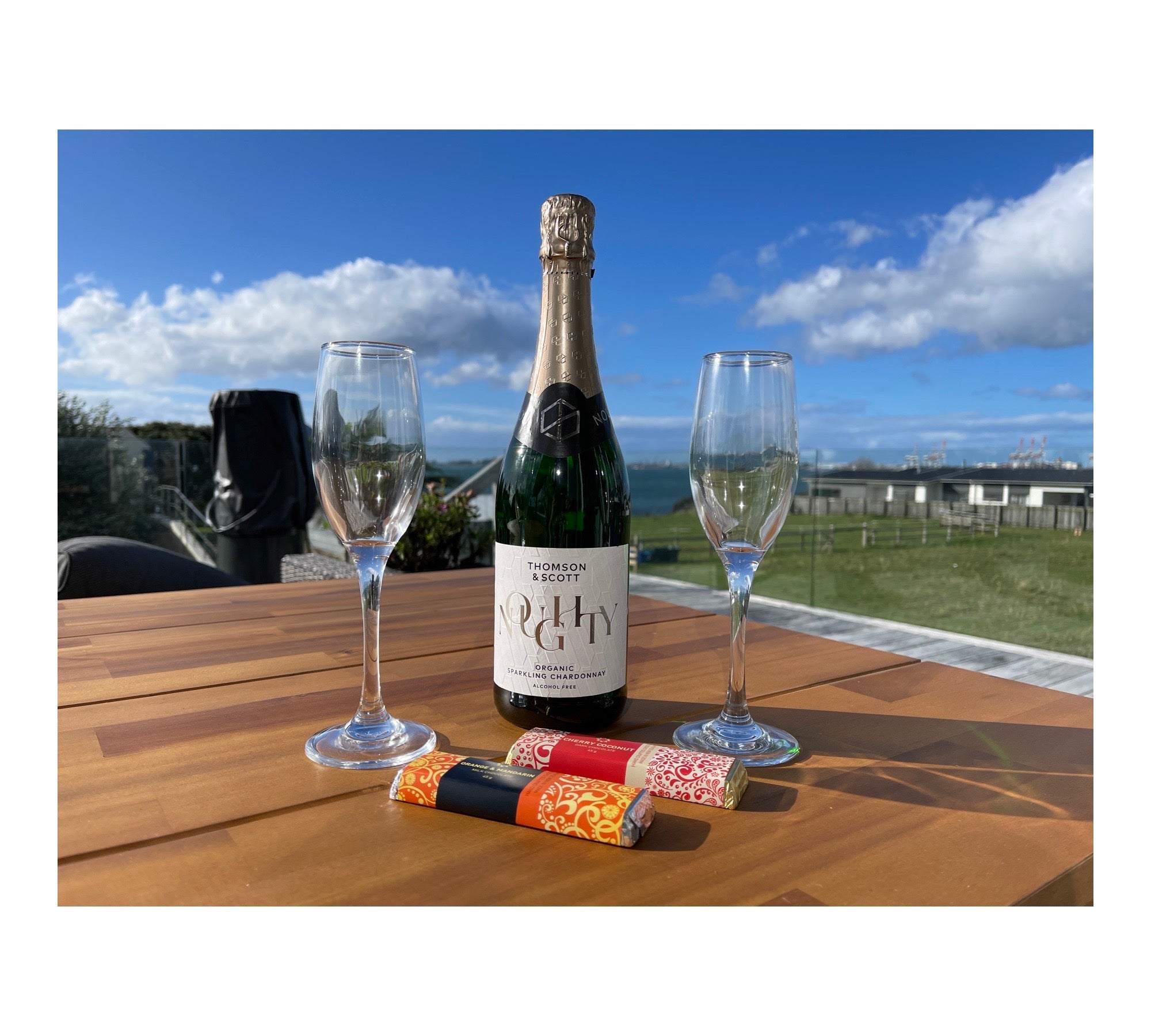A bottle of Celebrate alcohol-free sparkling wine from giftbox co. with two champagne flutes and party crackers on a wooden table outdoors, with a clear blue sky and green landscape in the background.
