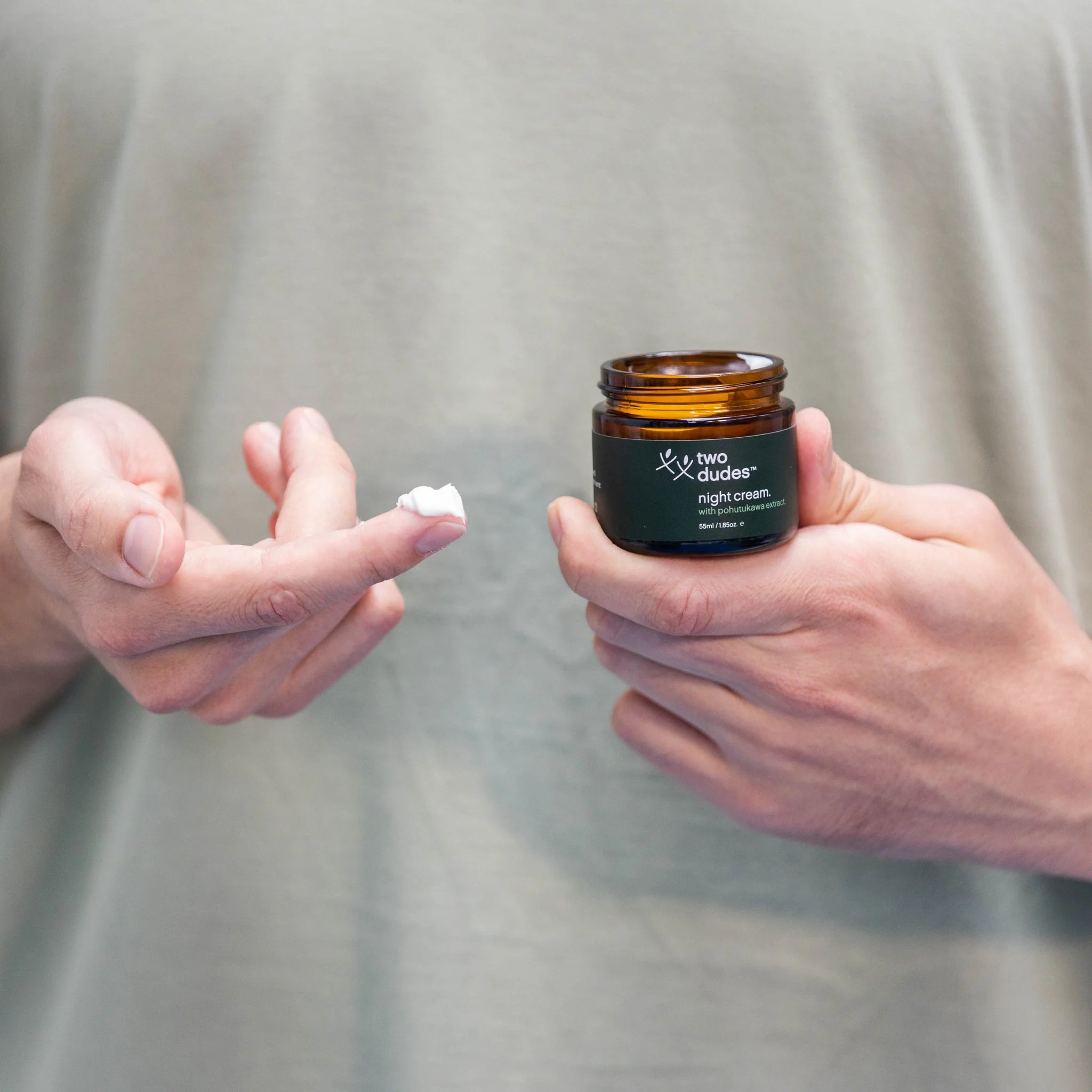 A person applying a dab of Night Cream by Two Dudes from a small jar labeled "Two Dudes" onto their fingertip.