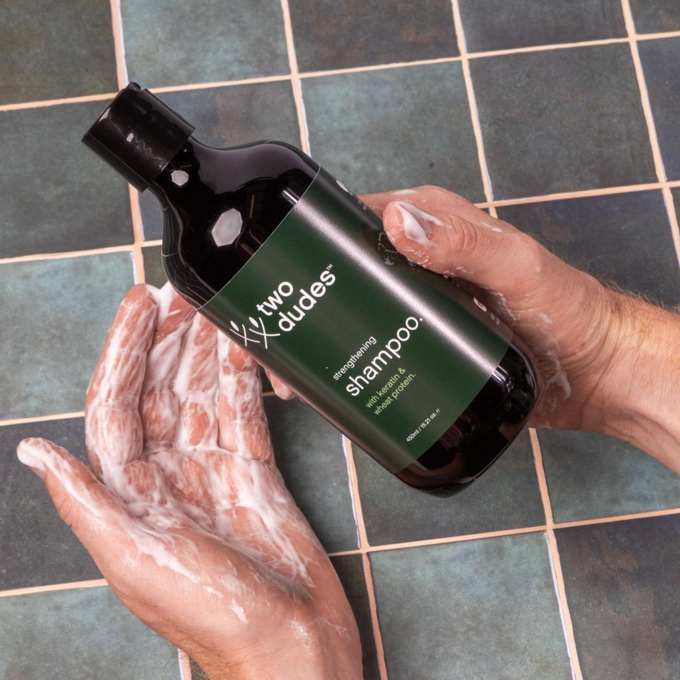 Two hands holding a bottle of "His" shampoo by giftbox co. over a tiled sink, with soapy water and suds visible. The setting suggests the shampoo is part of men's grooming products.