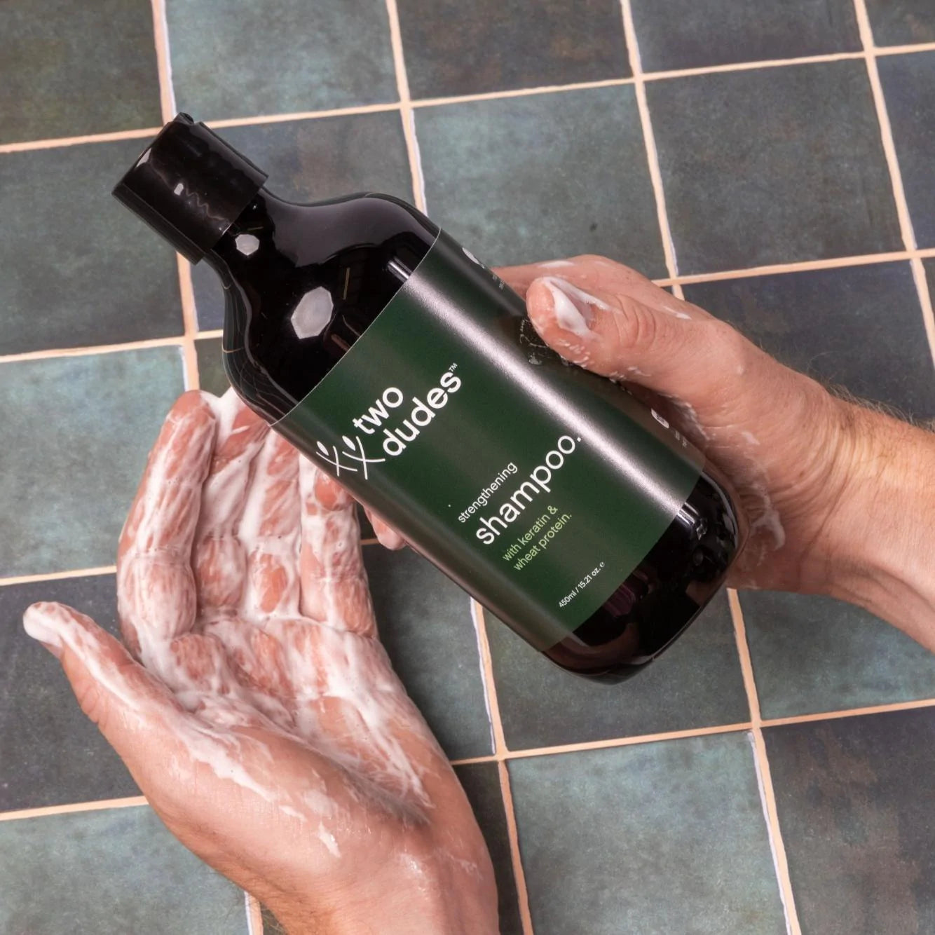 A person holding a black bottle of Two Dudes hair growth shampoo above their foamy, soaped hands, with a tiled surface in the background.