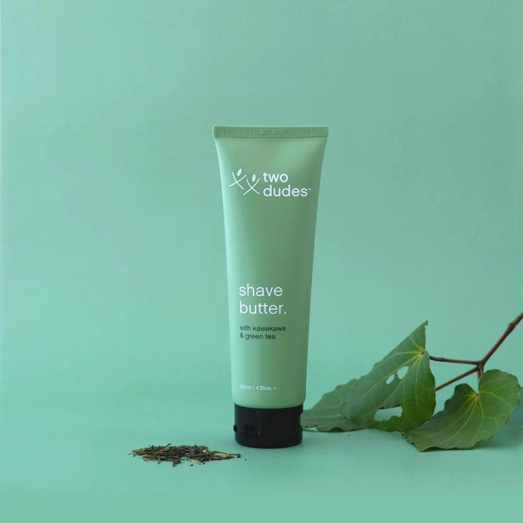 A tube of 'Two Dudes Shave Butter' with kowakawa & green tea, placed against a calm seafoam green background, accompanied by a scattering of leaves and small botanical elements nearby.
