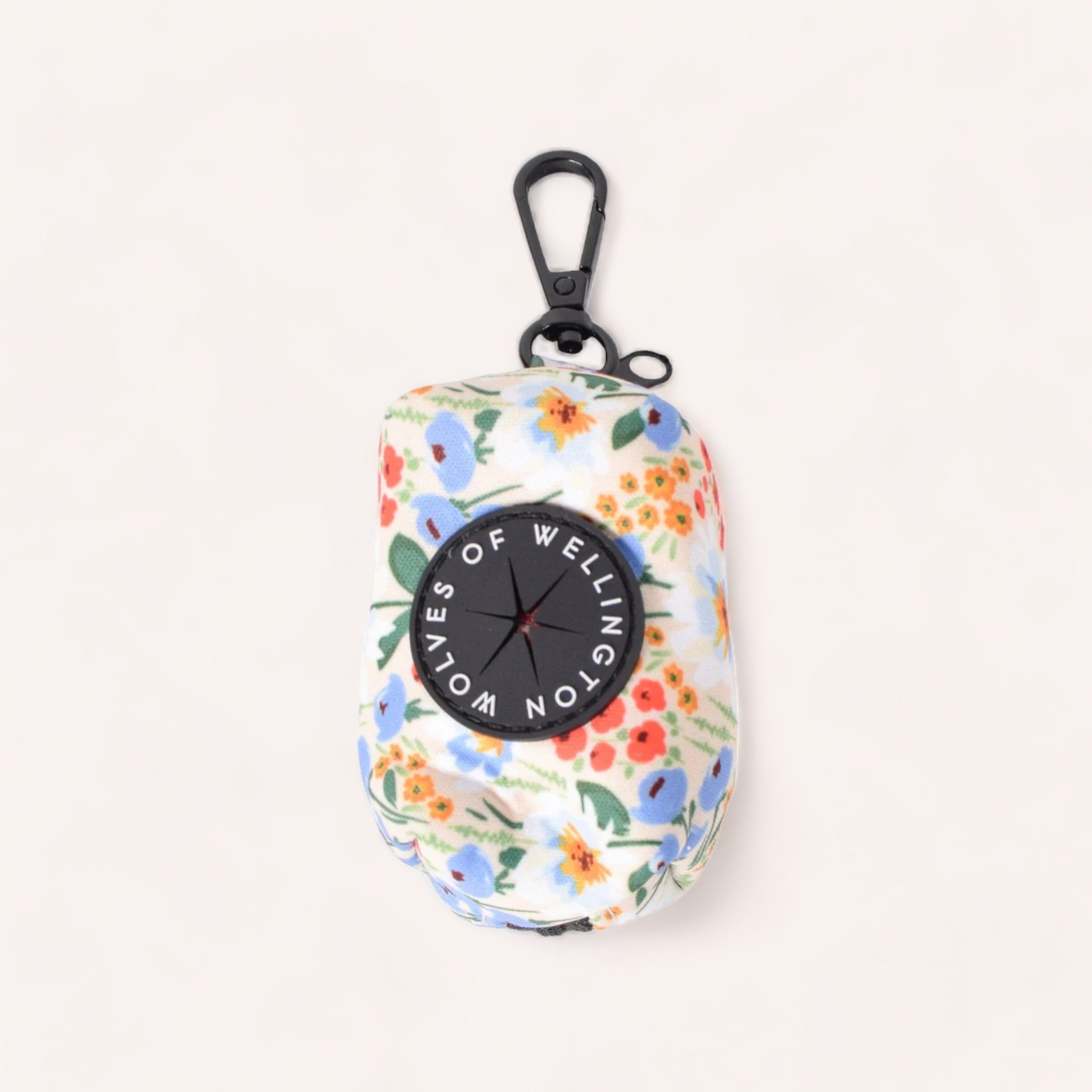 A photo of the Poop Pouch by Wolves of Wellington, featuring a floral-patterned design with a black carabiner at the top, perfect for dog parents to carry essentials like dog poop bags. The pouch is predominantly white with colorful flowers in shades of blue, orange, and green. A black circular tag on the front reads "WELLINGTON LOVES OF." The pouch is set against a plain, light-colored background.