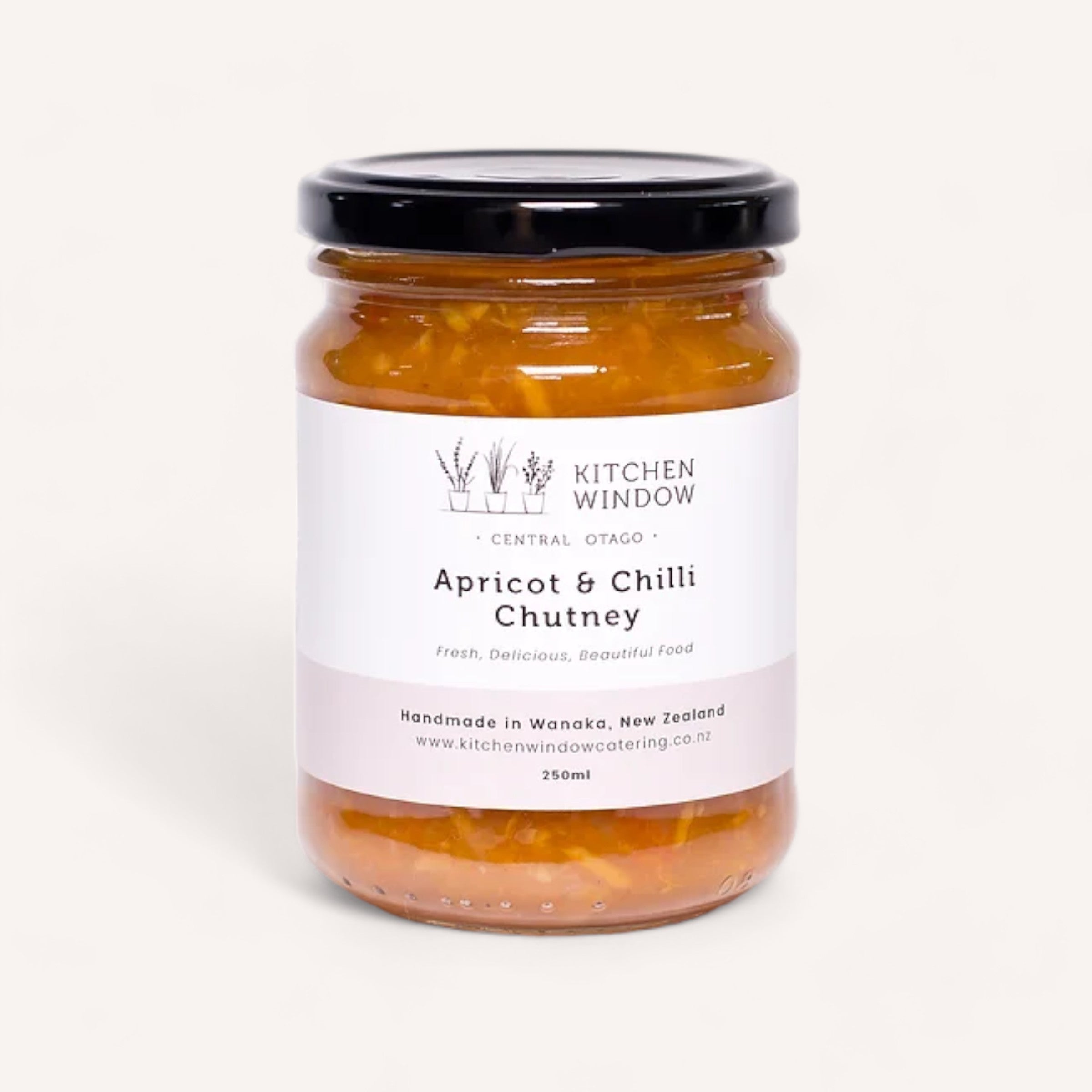 A 250ml jar of Kitchen Window's Apricot & Chilli Chutney, adorned with a black lid, features a minimalist label that reads "Fresh, Delicious, Beautiful Food" and highlights its handmade origin in Wanaka, New Zealand. This chunky orange chutney pairs wonderfully with creamy cheeses.