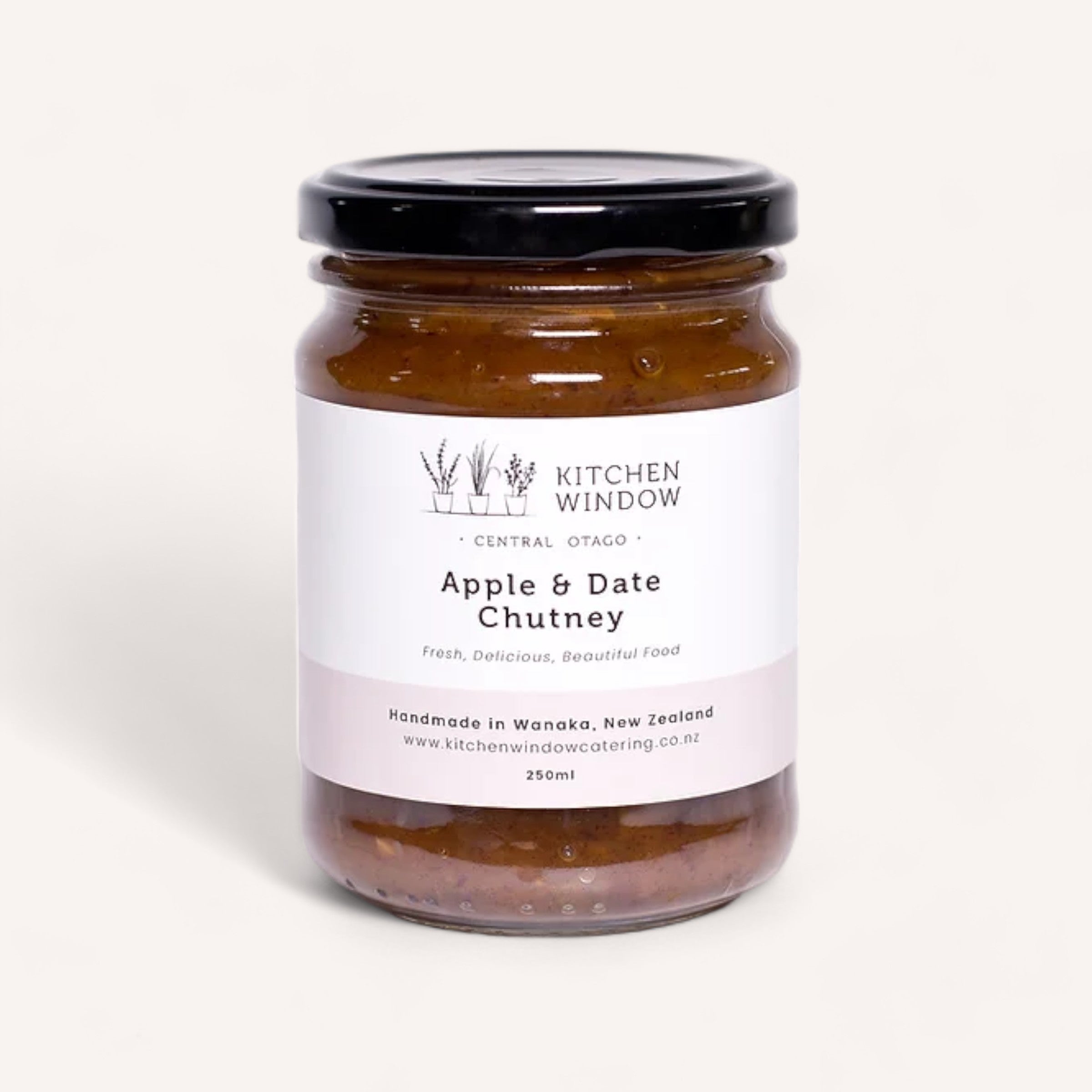 A jar of Apple & Date Chutney by Kitchen Window, known for its sweet and spicy flavor, is showcased against a white background. It has a black lid and a label adorned with botanical illustrations. The label highlights it as an award-winning preserve, handmade in Wanaka, New Zealand, with a 250ml volume.