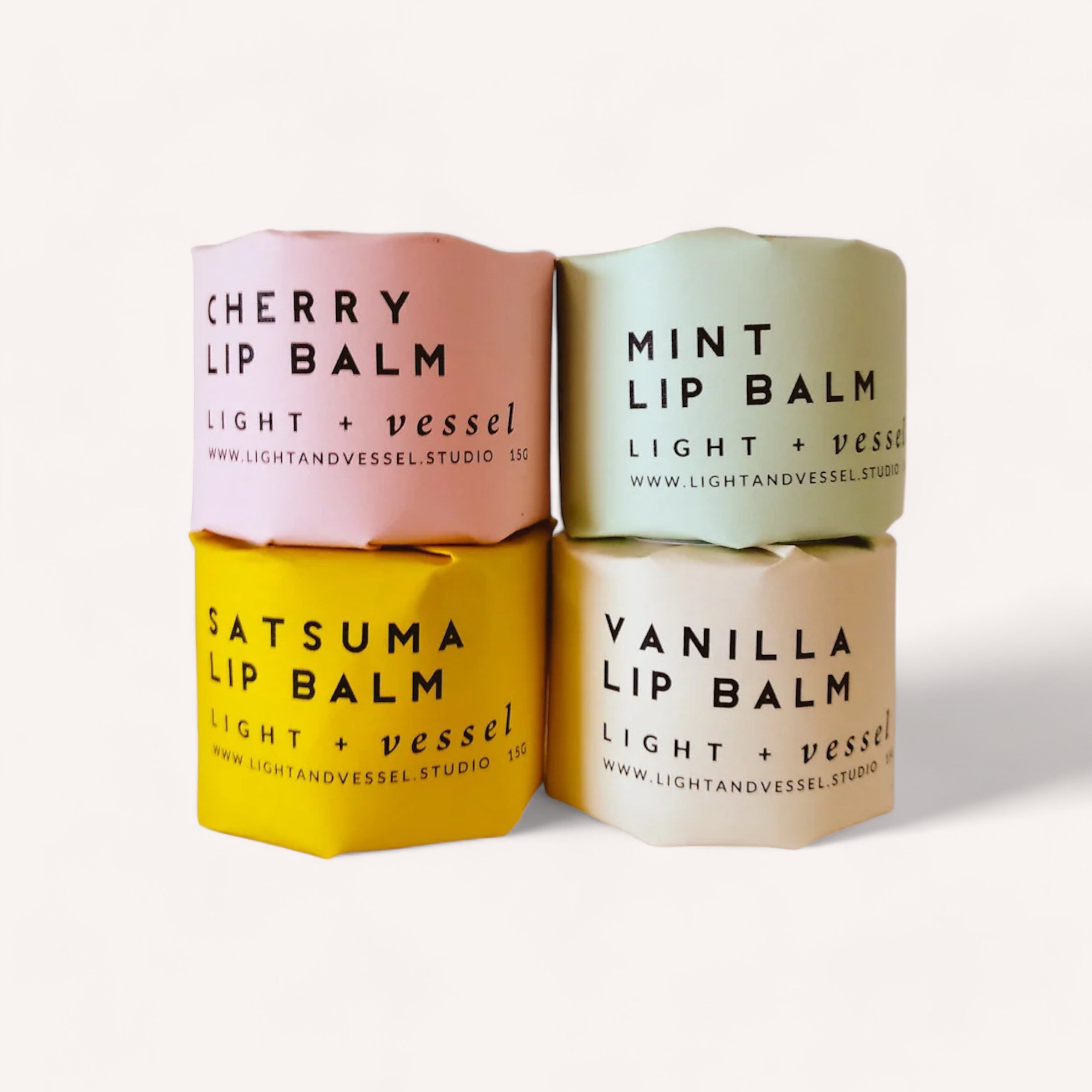 Four colorful Lip Balm by Light + Vessel containers in a row, each featuring a different organic flavour oil: cherry, mint, satsuma, and vanilla.