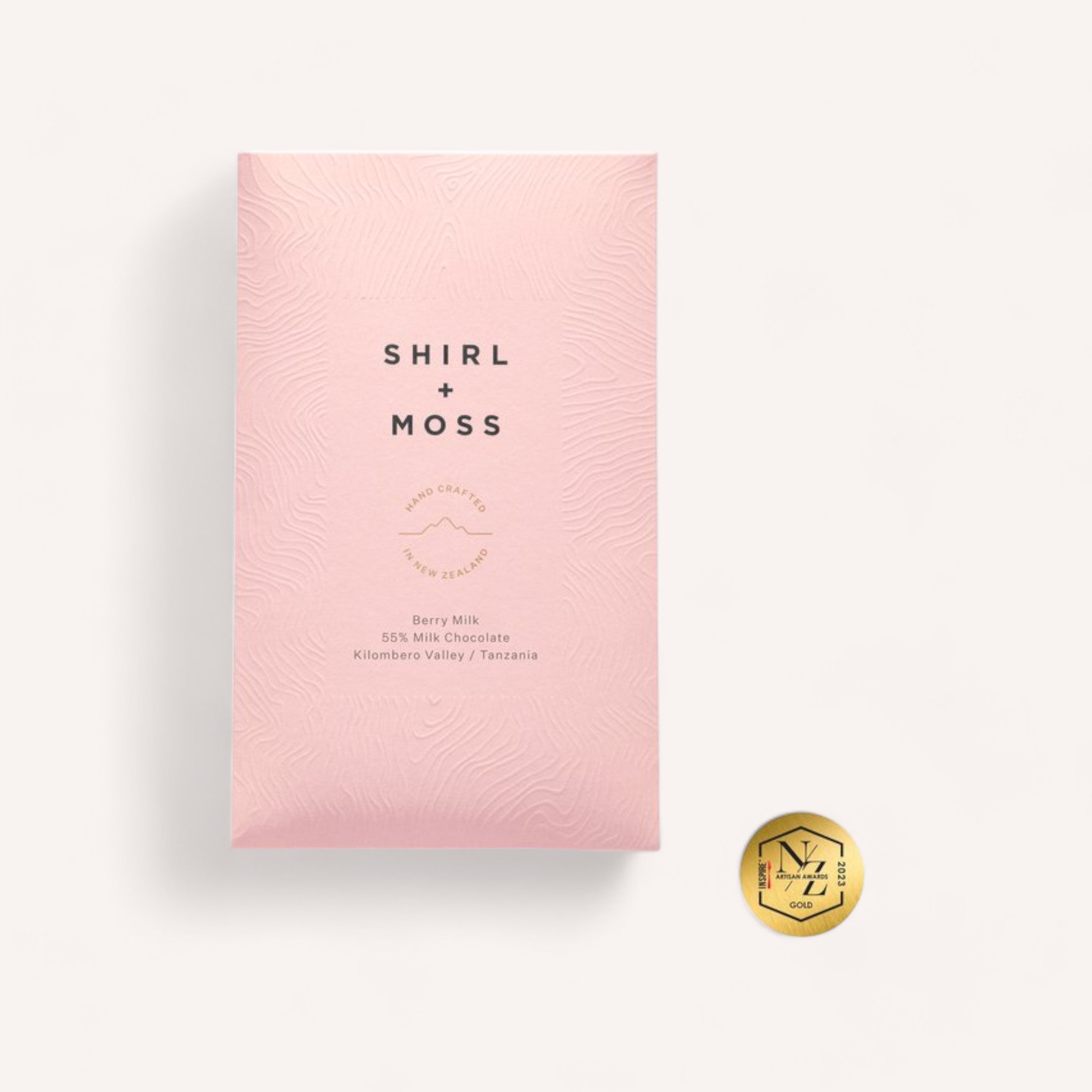 The image displays a pink wrapper for the Chocolate Bar by Shirl & Moss. The packaging is adorned with a wavy line texture and the brand name "Shirl & Moss" in black. It also includes information about their Berry Milk Chocolate, specifying "55% Milk Chocolate, Kilombero Valley, Tanzania." Additionally, a small gold seal signifies the award-winning quality of the chocolate next to the wrapper.