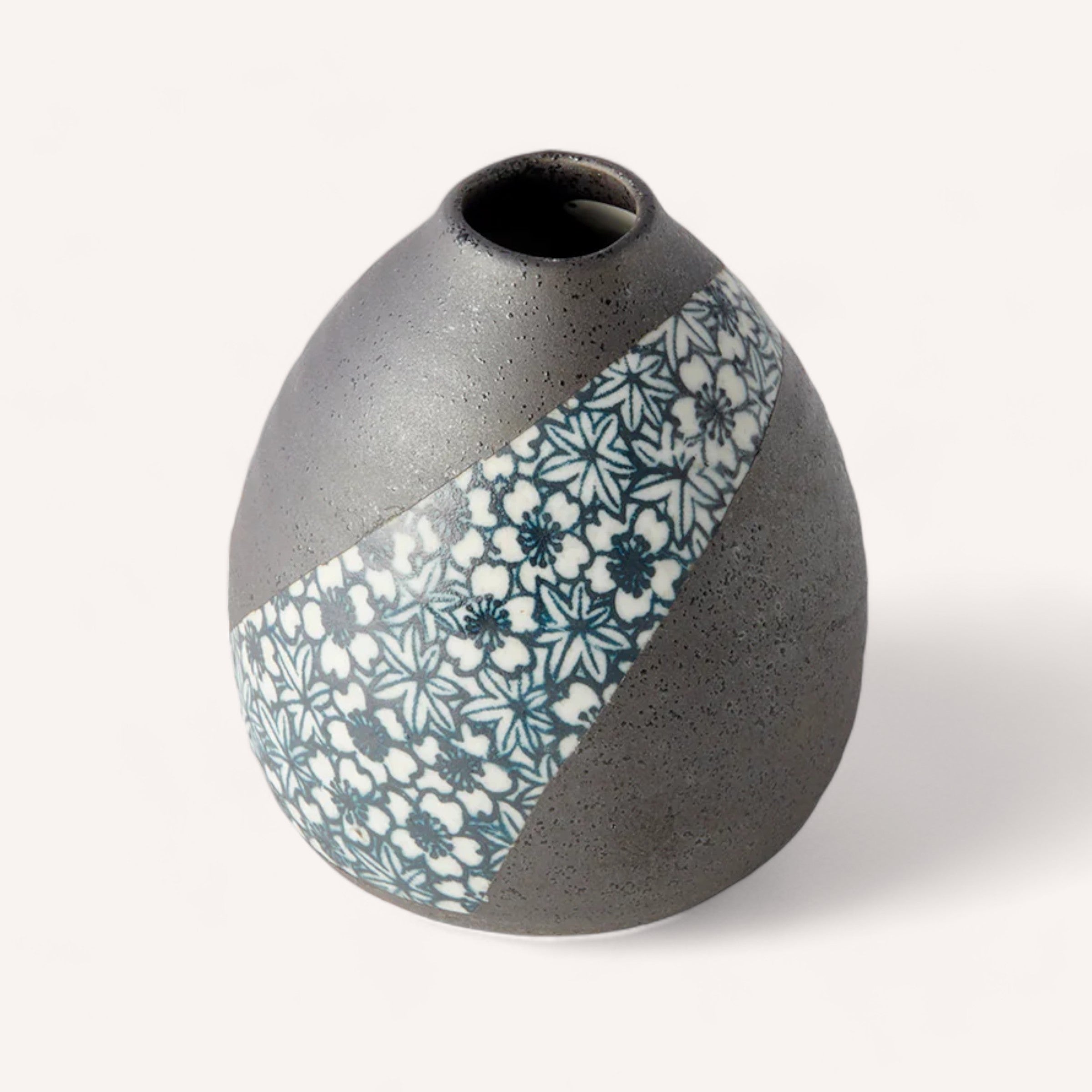 The Sakura Teardrop Vase by Naibu is a small, round, gray ceramic piece with a narrow opening, ideal for displaying single flowers. Handcrafted in Japan, it boasts a diagonal strip of black and white floral pattern over its slightly rough surface, creating an elegant contrast against a plain white background.