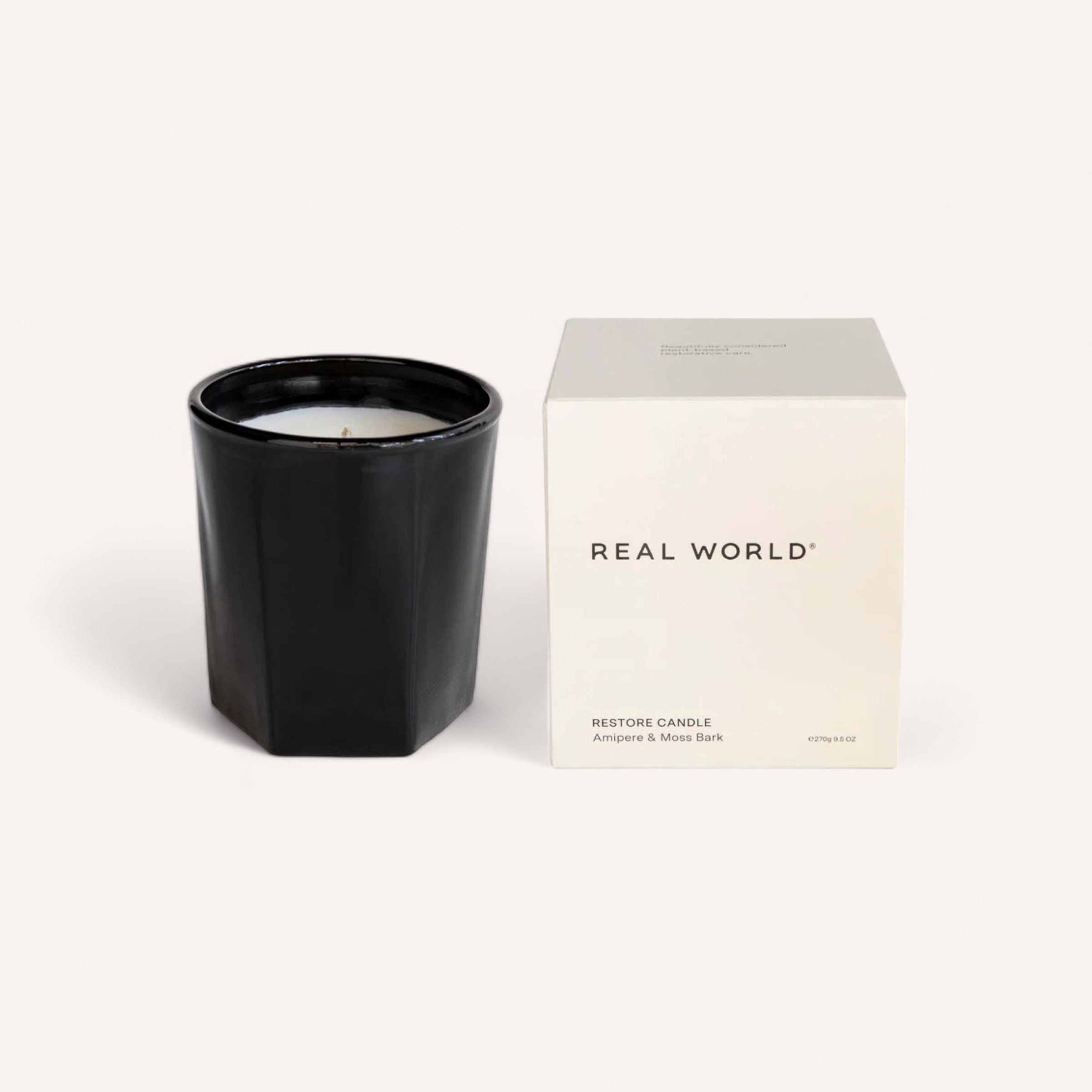 A black scented Amipere & Moss Bark candle by Real World next to its minimalist packaging with the label "real world, restore candle, amber & moss bark," a sustainable choice.