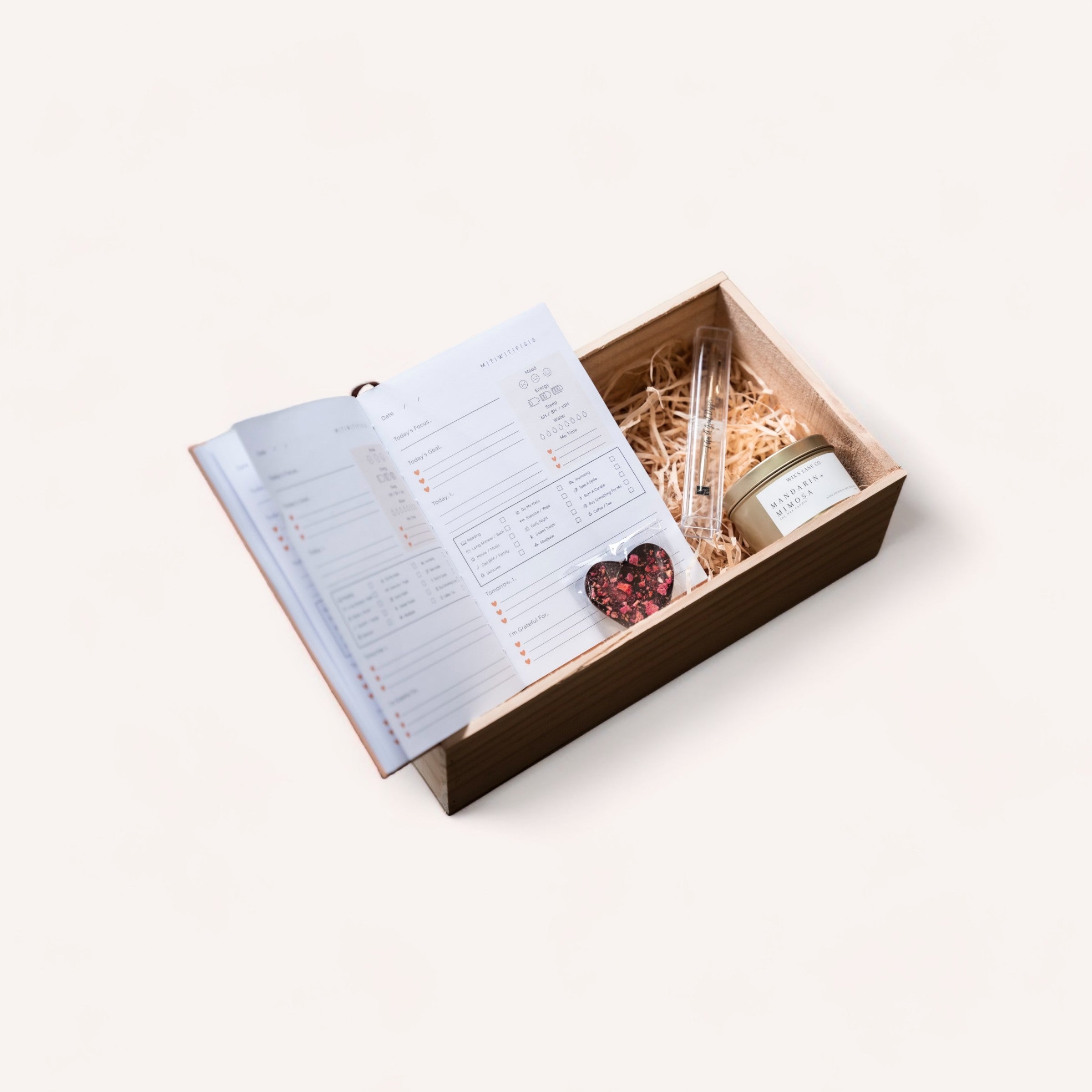 A minimalist presentation of an open Mindful Moments self-care gift box with a reflection journal on top and a tranquil candle tucked inside amidst protective straw padding, all against a clean, white background.