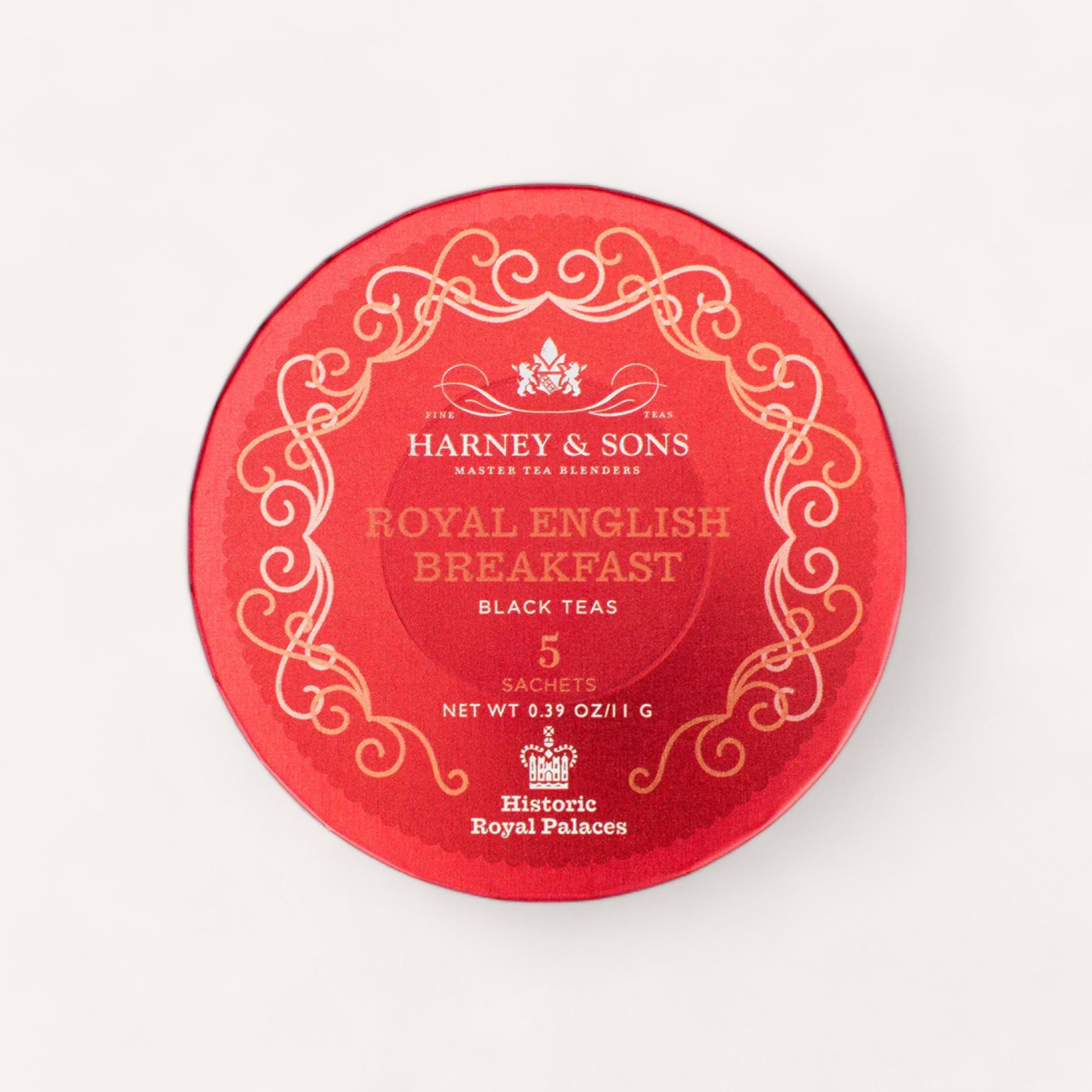 A round, red tin featuring the Tea Travel Pack by Harney & Sons, embellished with intricate gold patterns and text, contains pyramid silken sachets of Royal English Breakfast black tea. The pack includes 5 sachets with a total net weight of 0.39 oz (11 g). The bottom text reads "Historic Royal Palaces.