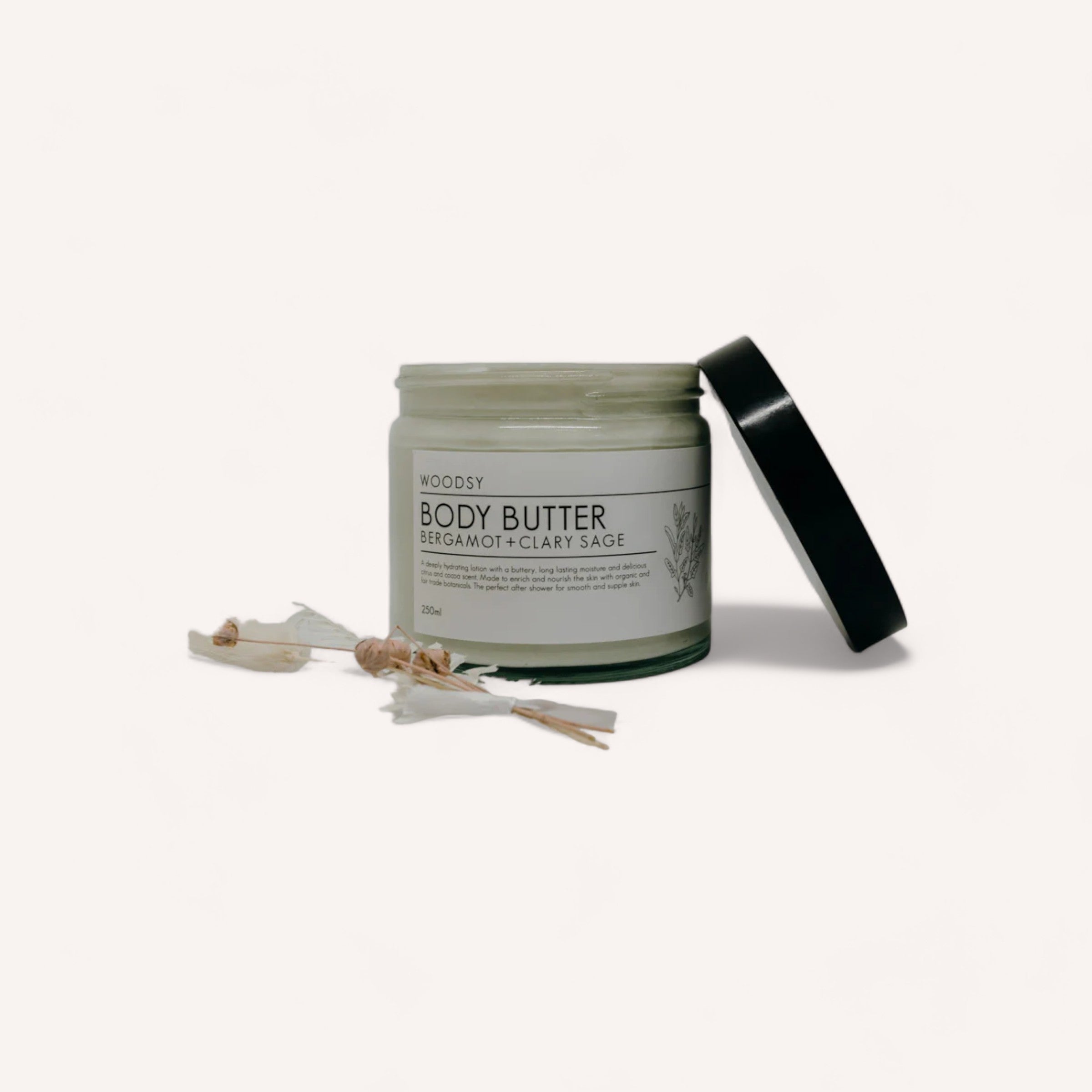 Sentence with replaced product: A jar of organic skin care Sage & Bergamot body butter by Woodsy Botanics, alongside a small bundle of dried botanicals, on a plain white background.