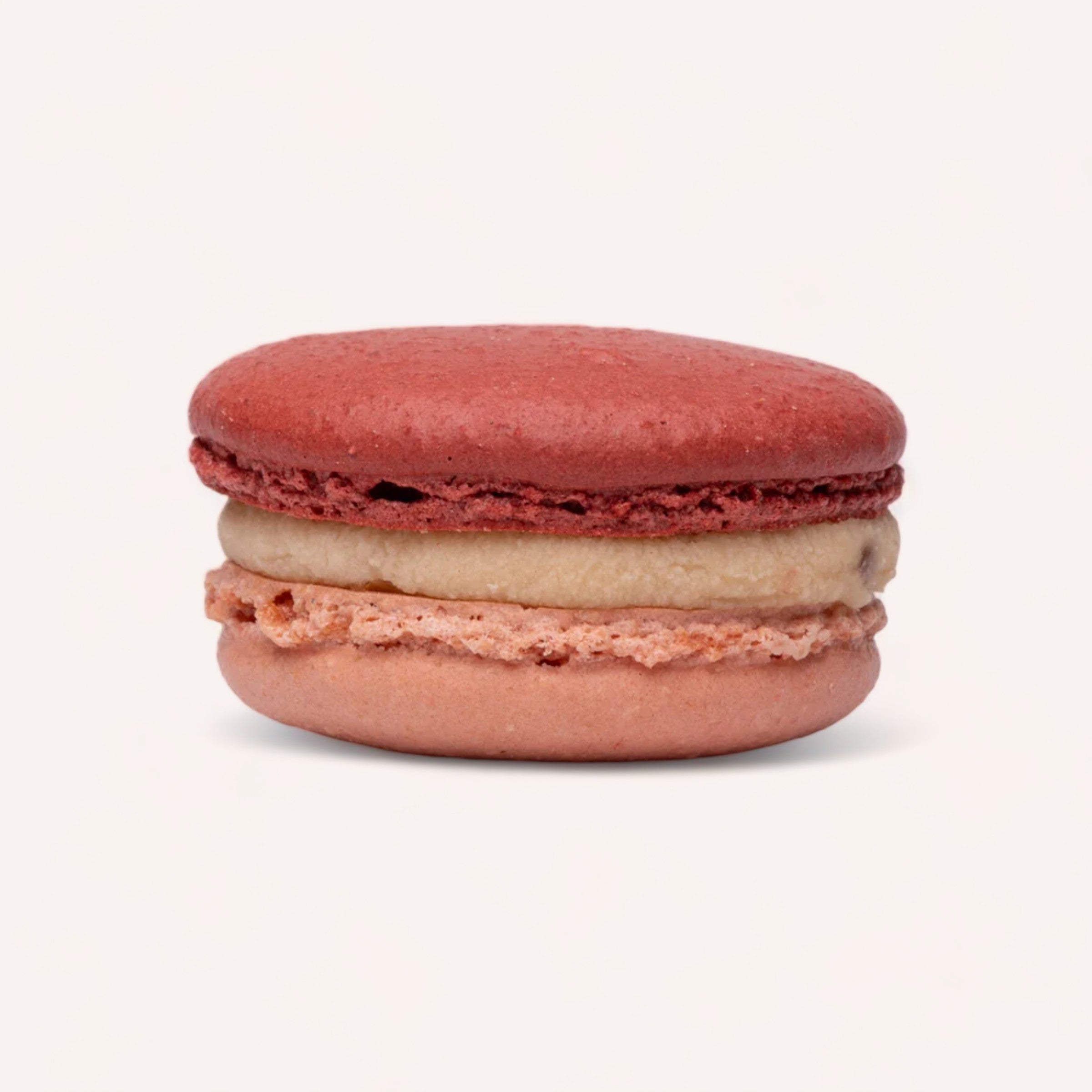 A solitary macaron from J'aime les Macarons is showcased against a white background, featuring a smooth red top and bottom shell with a creamy, light brown filling. This macaron is handmade in Christchurch using the finest ingredients, highlighting its texture and vibrant color contrast.