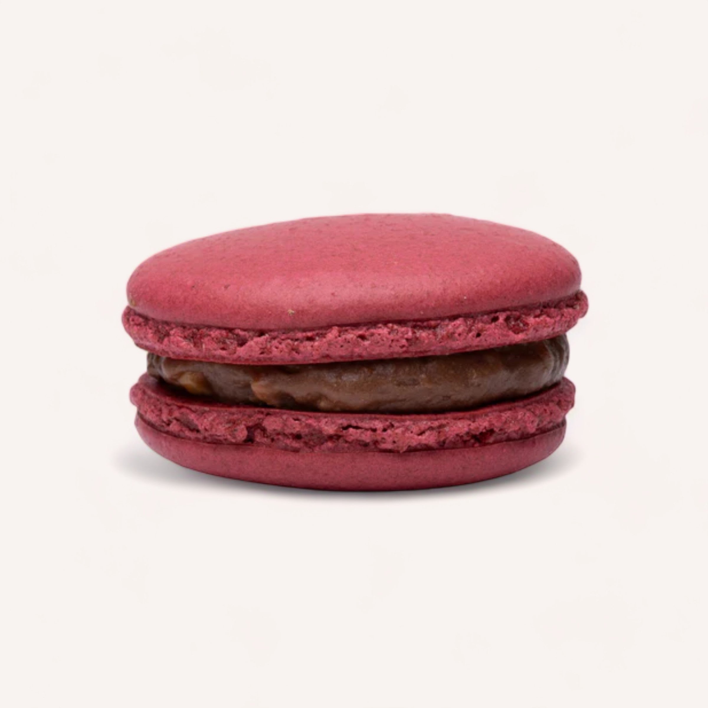 A single macaron from J'aime les Macarons, featuring a pinkish-red hue and a smooth shell, filled with rich dark chocolate ganache, highlights its classic round shape on a plain white background. Handmade in Christchurch by J'aime les Macarons using the finest ingredients, this macaron appears crisp yet slightly chewy, with delicate ruffled "feet" at the base.