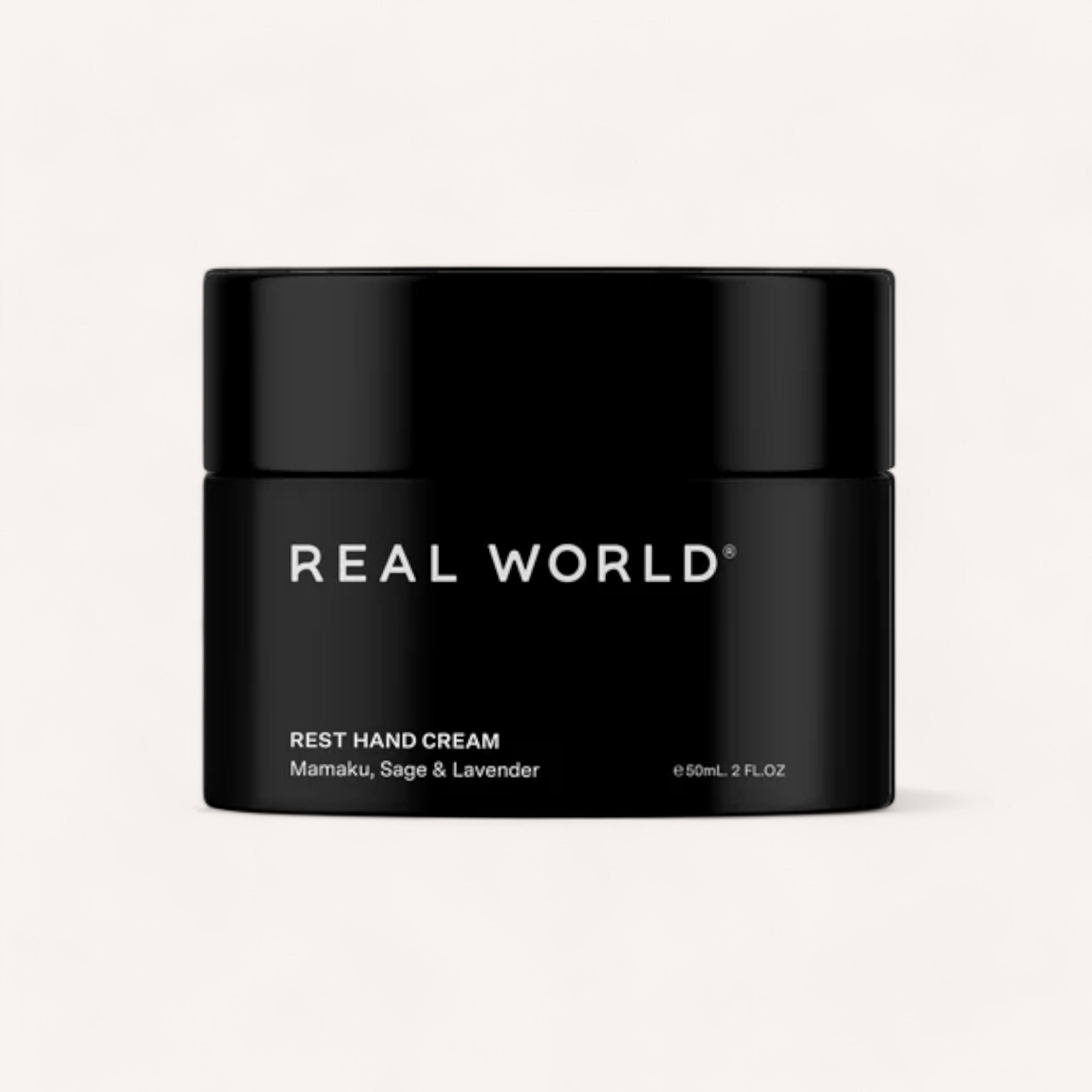 A sleek black jar with a matching black lid, labeled "REAL WORLD®" in white text. The product is "Hand Cream by Real World" containing Mamaku, Sage & Lavender. The size is indicated as 150mL or 2 FL.OZ. This New Zealand hand cream boasts a minimalist design and promises natural, restorative benefits.