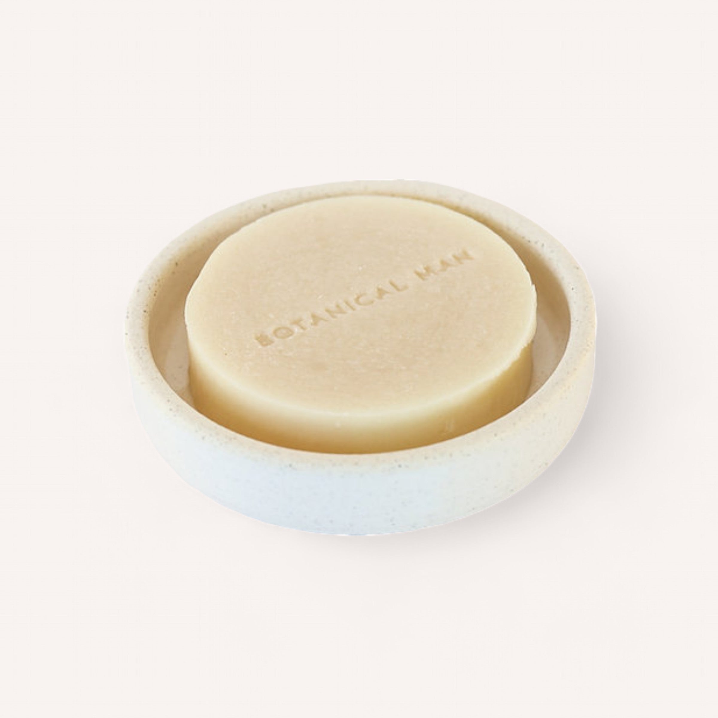 A round, beige Shaving Soap by Botanical infused with shea butter is placed in a matching white, round dish. The soap is embossed with the text "BOTANICAL MAN" on its top surface. The dish features smooth, slightly raised edges and showcases a simple, minimalist design. The background is a plain, light color.