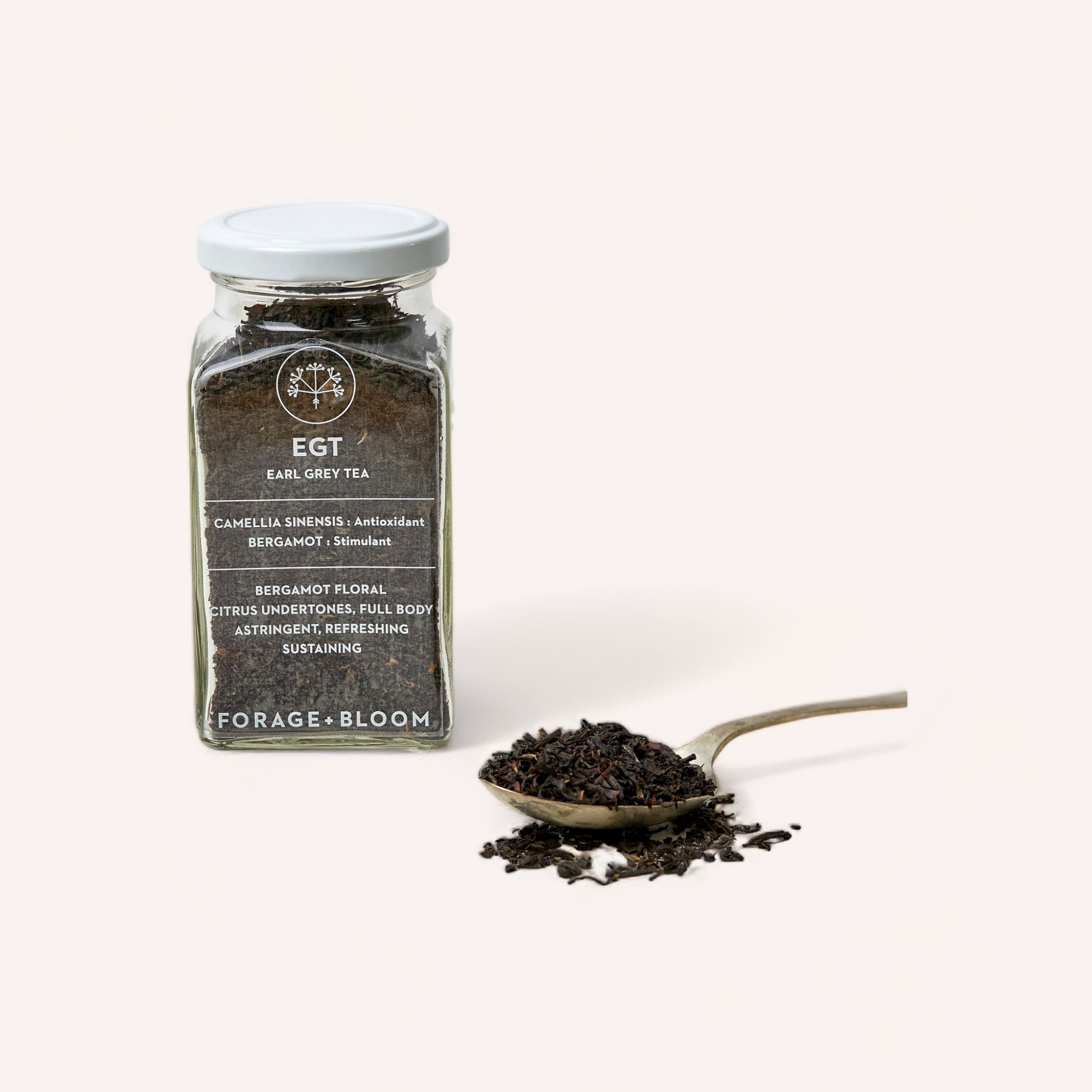 A jar of Forage + Bloom Earl Grey tea, infused with bergamot, alongside a teaspoon of loose tea leaves on a white background.