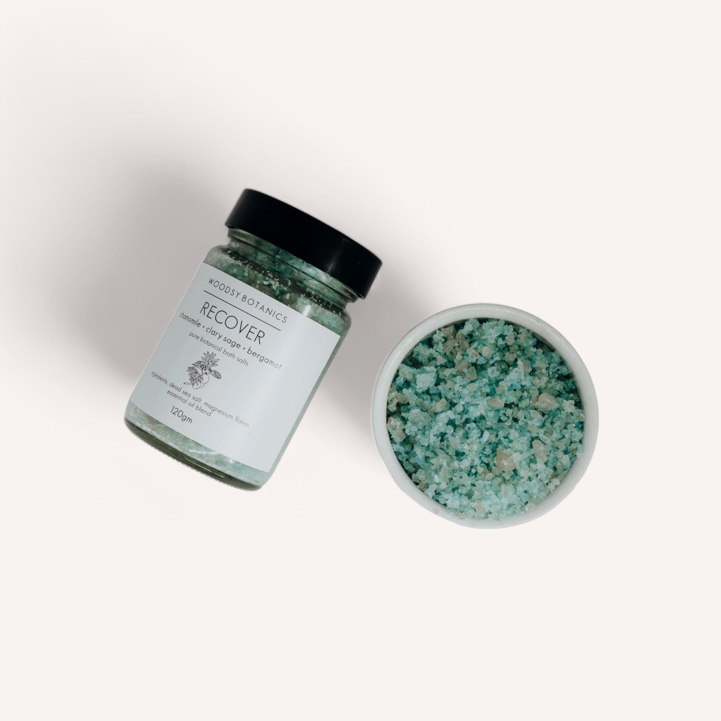 A jar of Woodsy Botanics' Bath Lovers Gift Set labeled "restore" alongside its open lid, revealing the green crystals inside, on a clean white background.
