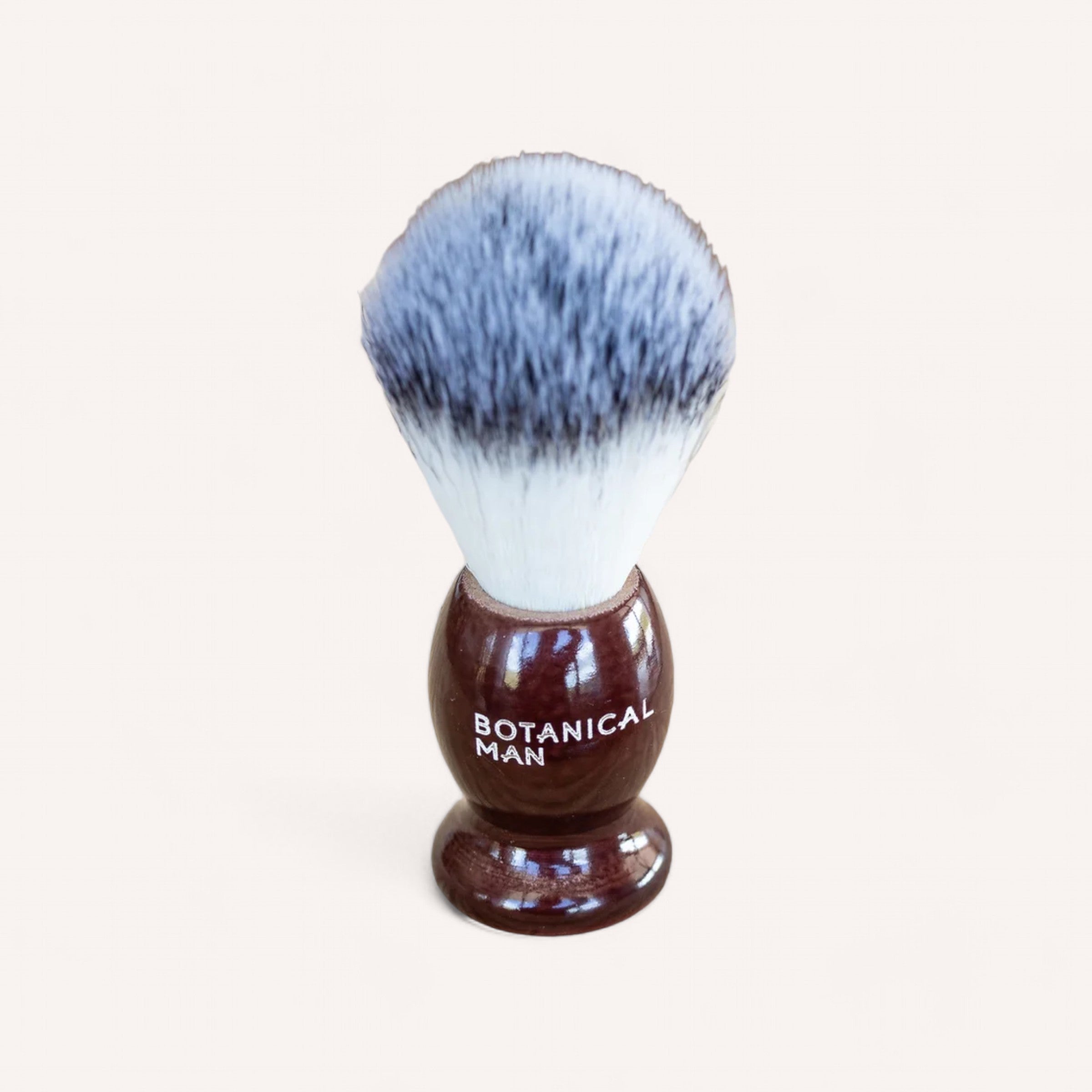 A shaving brush with white and dark gray bristles and a glossy dark brown handle. The bulbous base reads "BOTANICAL" in white text. Standing upright against a plain white background, this brush pairs perfectly with the Botanical Man Shaving Set for an elevated grooming experience from the renowned brand Botanical.