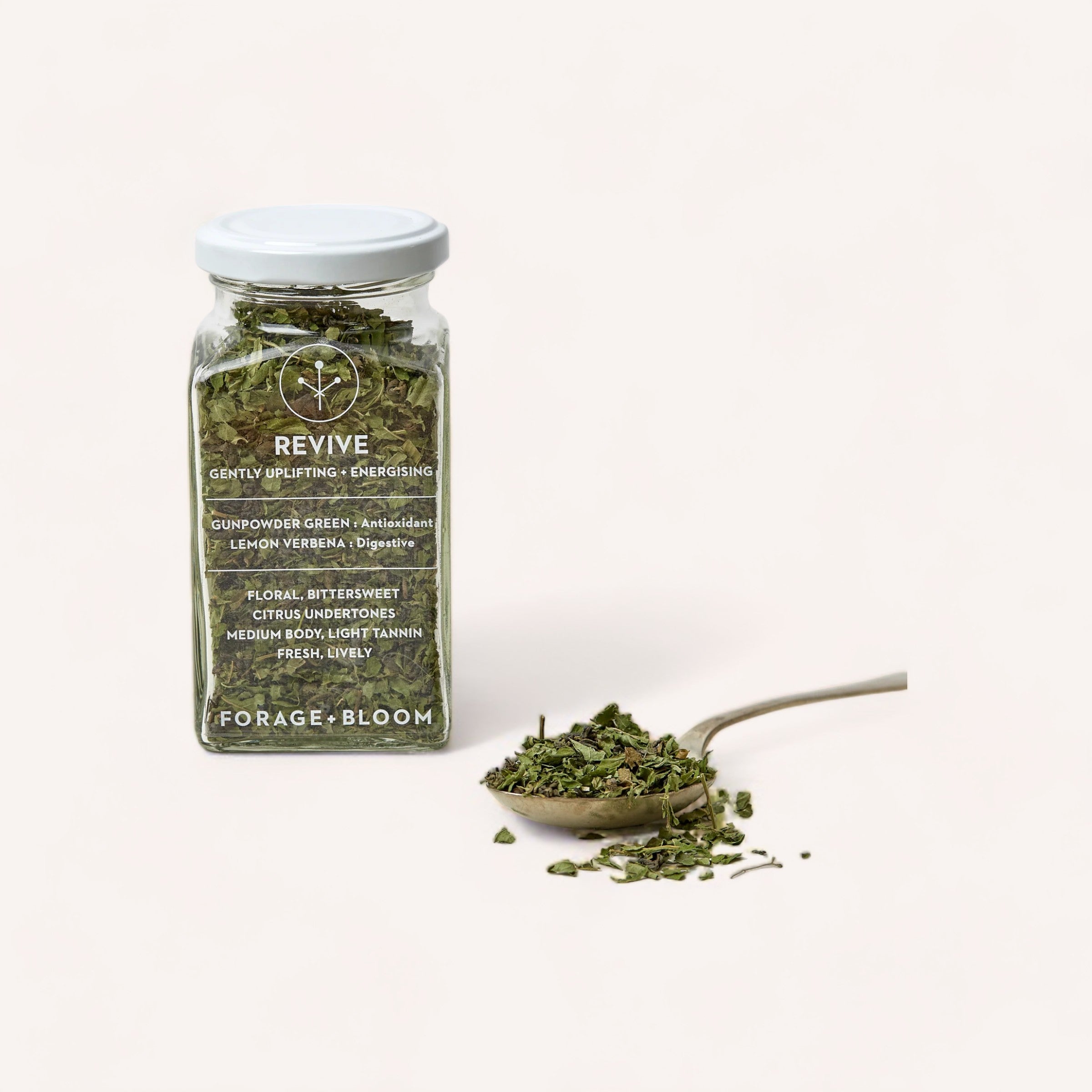 Jar of 'Revive Tea by Forage + Bloom' loose green tea leaves, known for its antioxidant properties, with a scattered pile of leaves beside it on a white background.