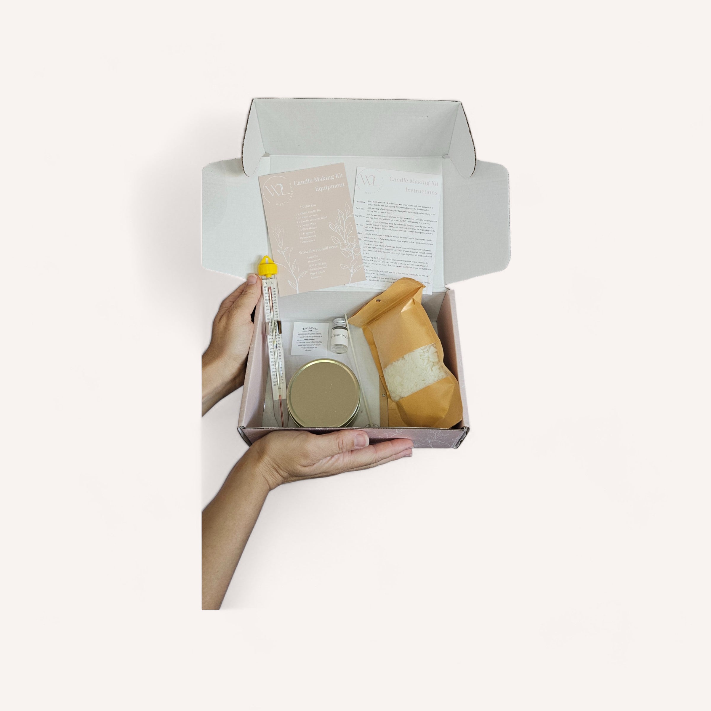 A person holding an open box filled with an assortment of skincare products and a Candle Making Kit by Wix's Lane Co against a white background.