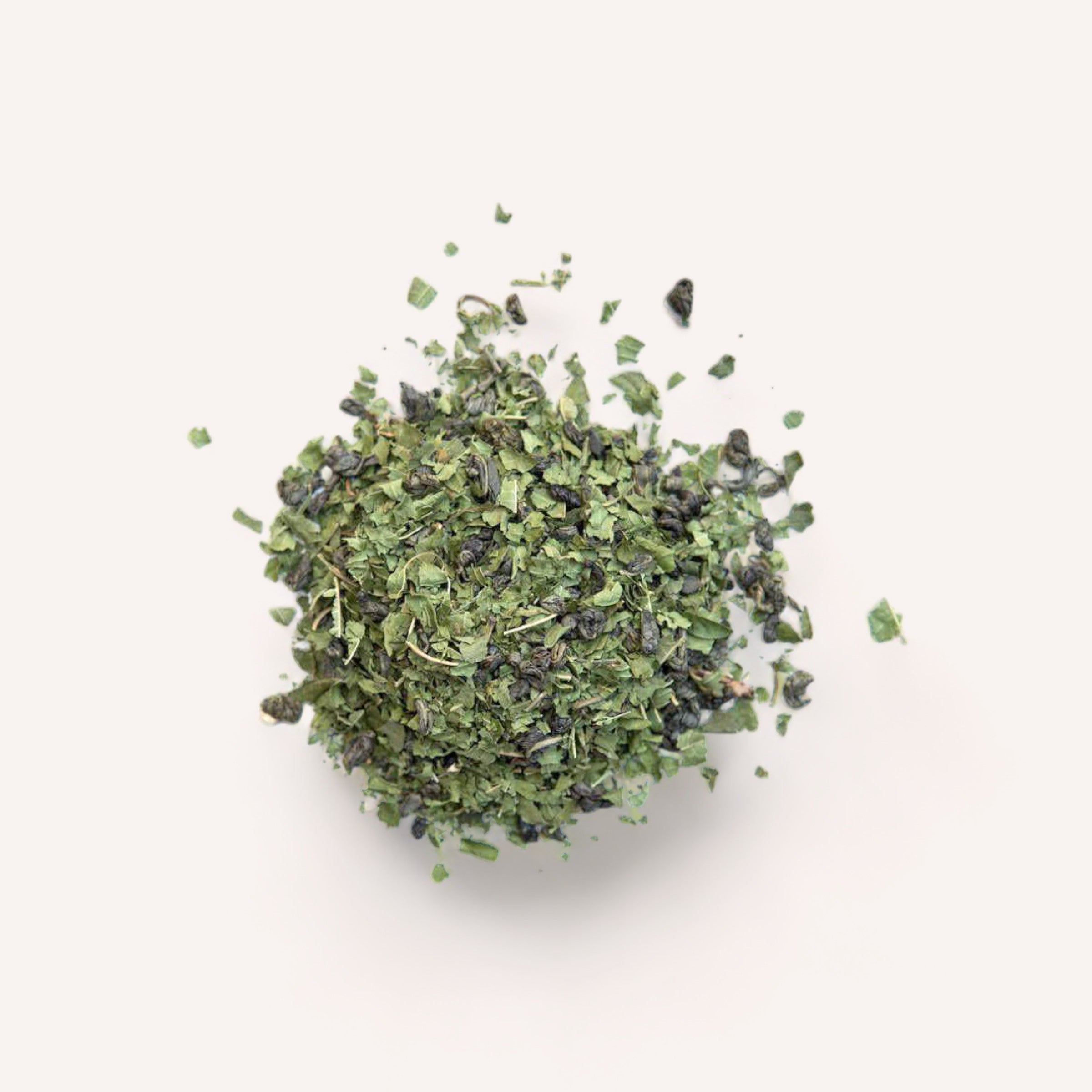 The *Revive Tea by Forage + Bloom*, a mixture of dried green tea leaves and herbs renowned for enhancing metabolism, is spread out against a simple white backdrop. This blend features small, curled leaves mixed with fine herbal pieces, presenting an organic look. Its vibrant green hue is rich in antioxidants.