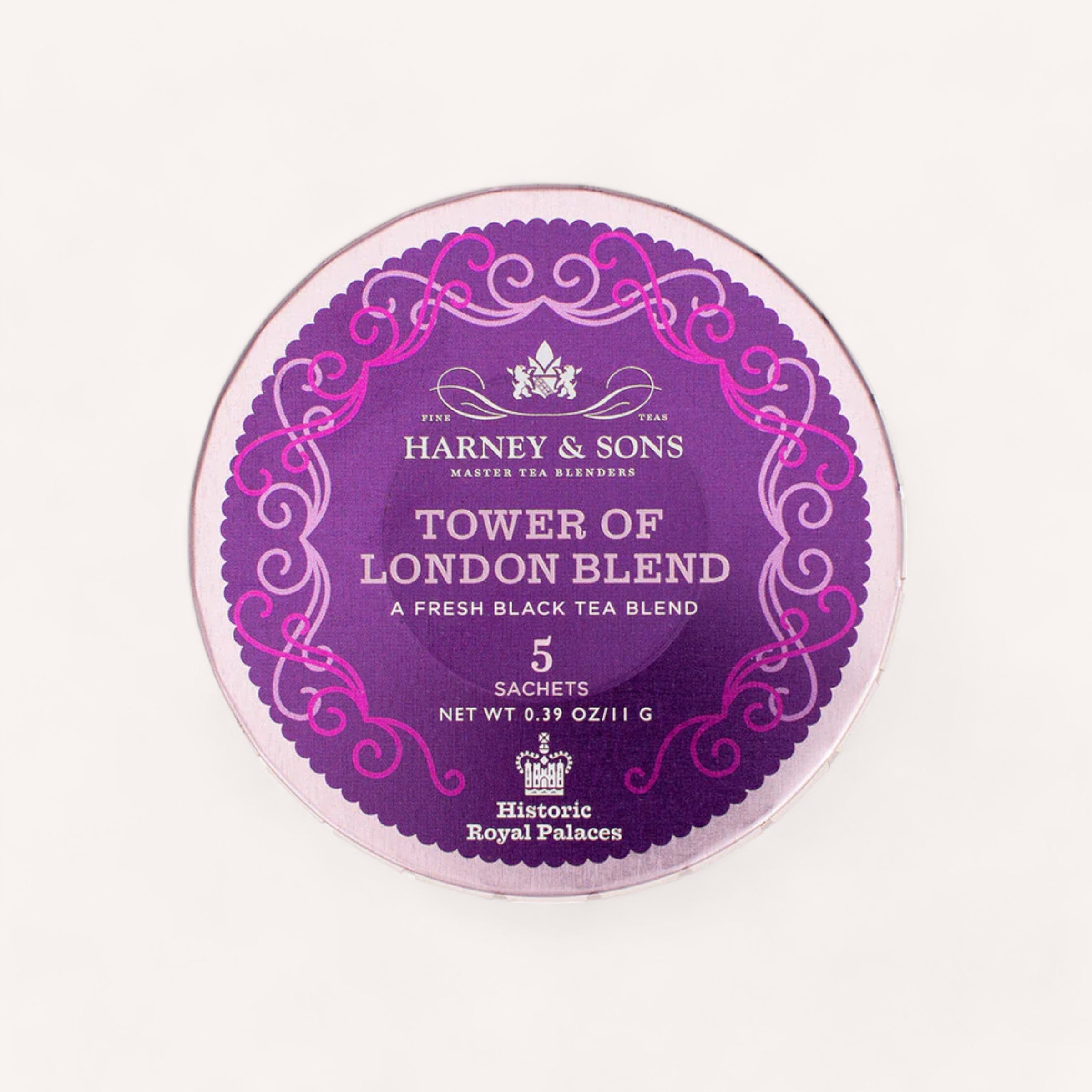 A round purple tea sachet package from Harney & Sons, similar to their sophisticated tea tins, showcases an elaborate pink border and logo at the top. It states "Tower of London Blend" with a description "A Fresh Black Tea Blend" and notes "5 pyramid silken sachets, net wt 0.39 oz/11 g," as well as "Historic Royal Palaces.