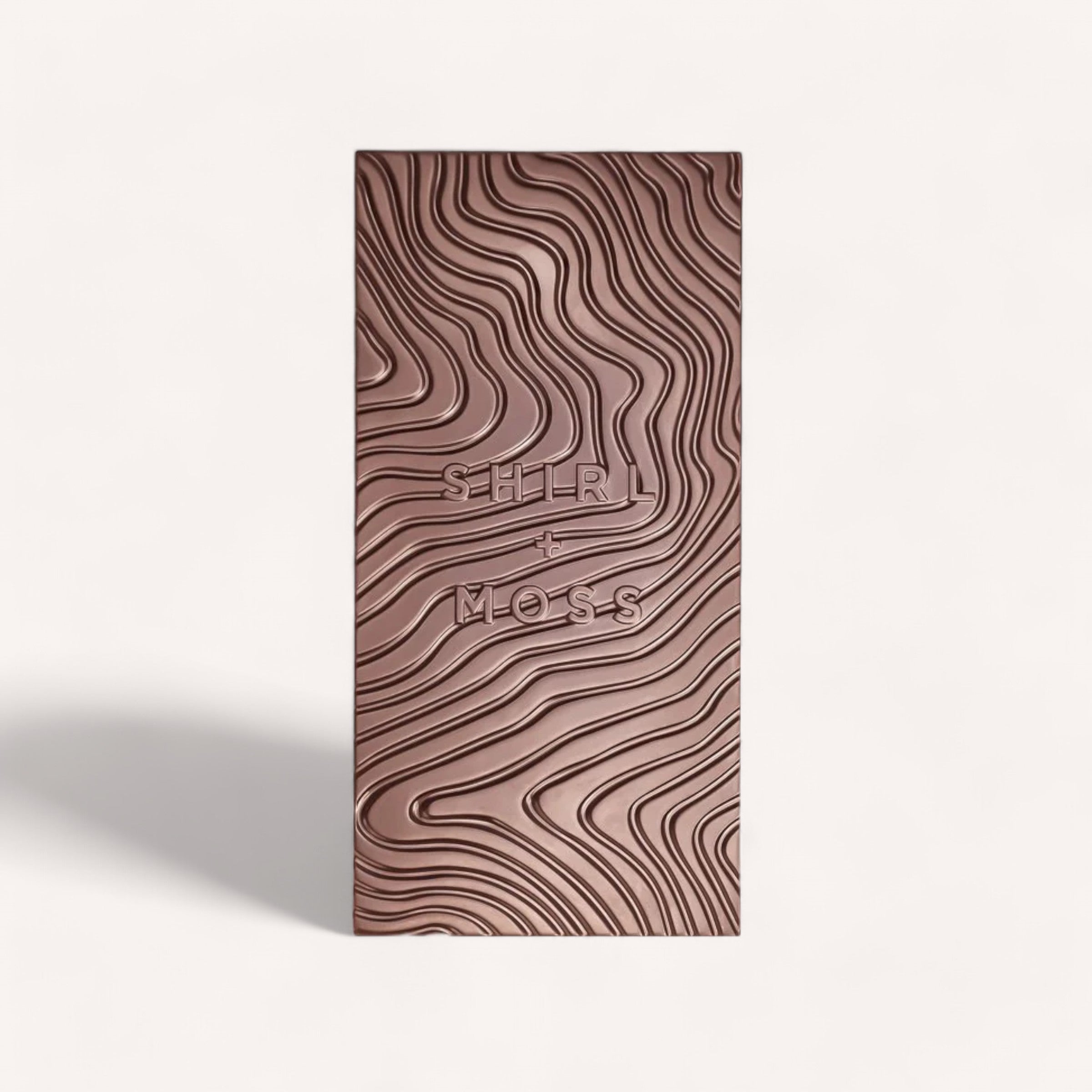 A rectangular bar of creamy milk chocolate with a textured, wavy pattern covers the entire surface. The words "Shirl & Moss" are embossed in the center of the Chocolate Bar by Shirl & Moss. The background is a plain, light color. A faint shadow is cast to the left of the bar.