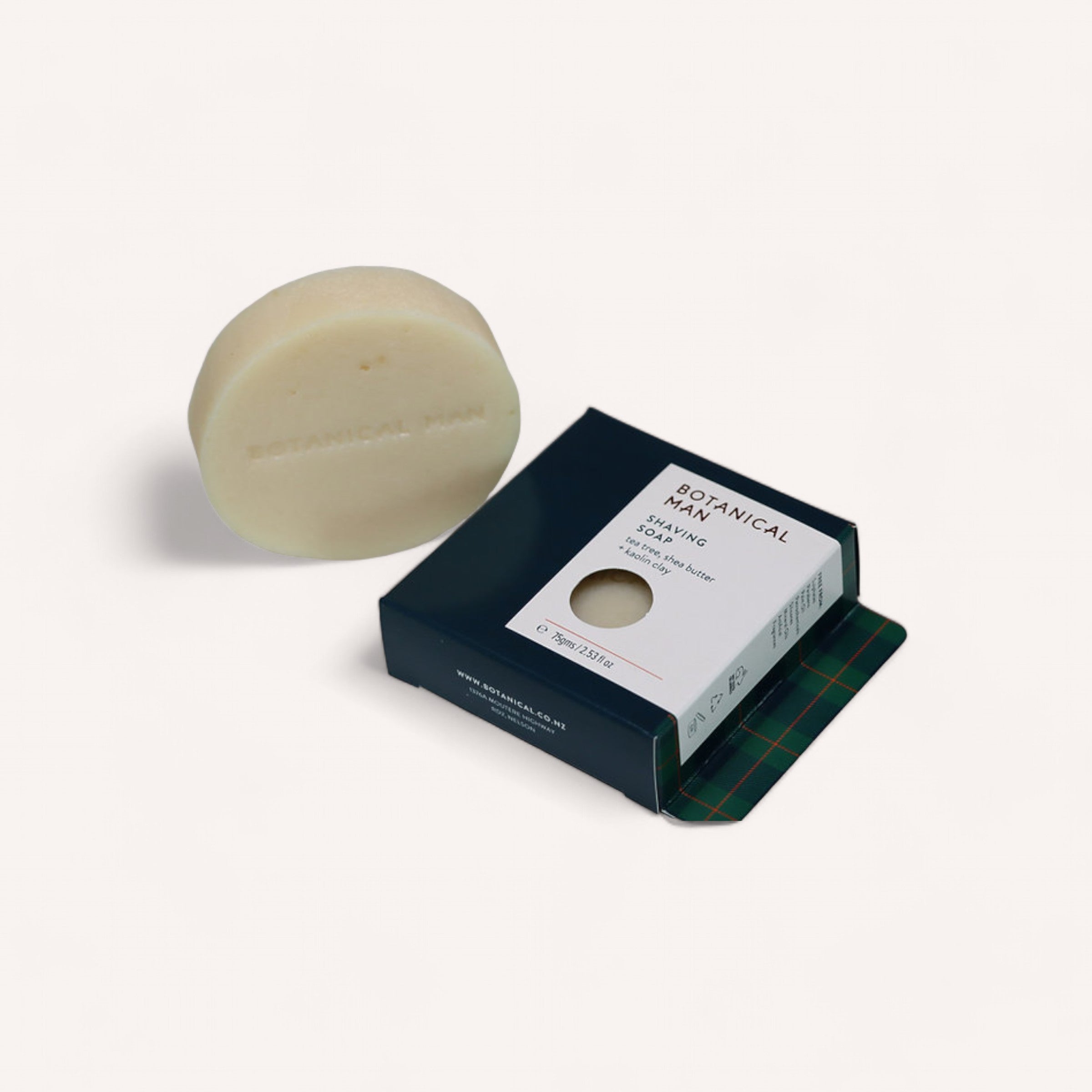 A round, beige shaving soap bar by Botanical is displayed next to an open black and white rectangular box labeled "Shaving Soap by Botanical." The soap, infused with tea tree and partially wrapped in dark green plaid paper, is visible through a circular cutout in the lid.