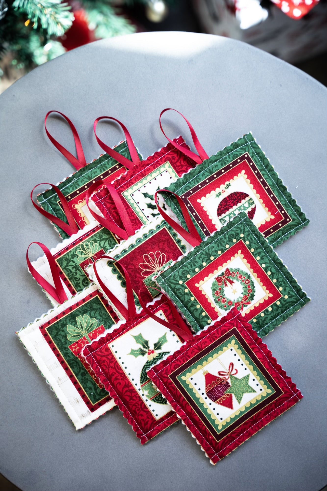 Introducing the Handmade Christmas Tree Decorations by giftbox co., a charming collection featuring intricate designs, each measuring 10cm square. These ornaments showcase festive motifs such as wreaths, stockings, and holly, beautifully framed in red, green, or white fabric. They come with red ribbon loops for easy hanging and are elegantly displayed on a gray circular surface.