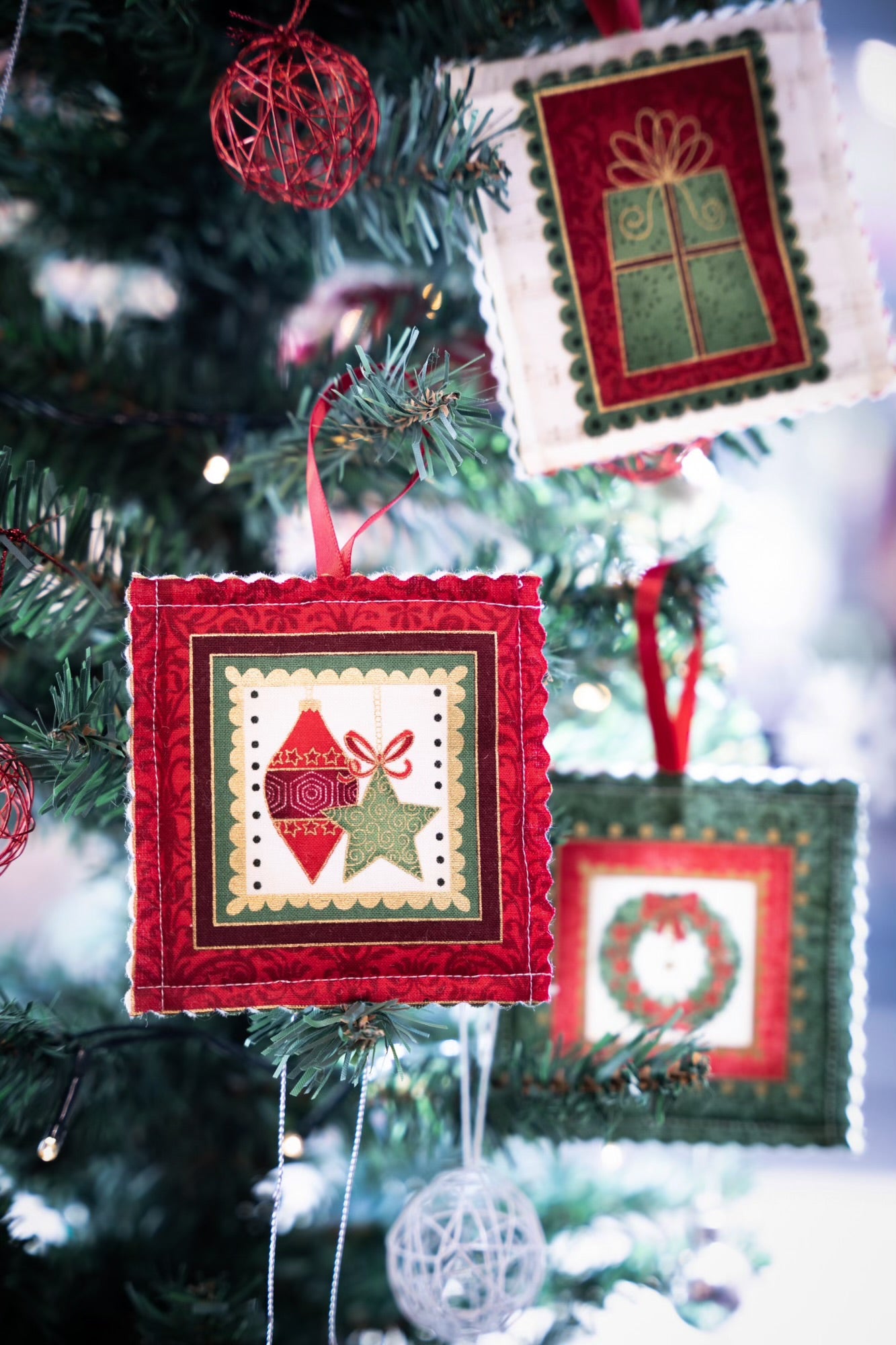 Close-up of a Christmas tree embellished with giftbox co.'s Handmade Christmas Tree Decorations, each measuring 10cm square. These quilted ornaments, showcasing designs like presents, hearts with stars, and wreaths, are highlighted by red ribbons. They dangle amid twinkling lights and round woven ornaments to create a cozy holiday atmosphere.