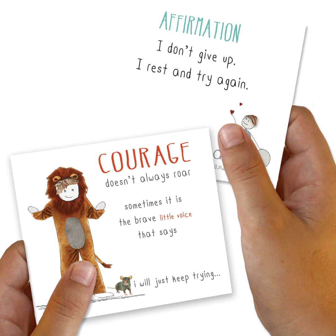 Close-up of hands holding two illustrated cards from the Positivity Pack by iCandy. The card in the left hand depicts a child in a lion costume with a smaller figure and text that reads: "COURAGE doesn't always roar - sometimes it is the brave little voice that says I will just keep trying." The card in the right hand reads: "AFFIRMATION - I don't give up. I rest and try again." A perfect blend of encouragement and resilience.