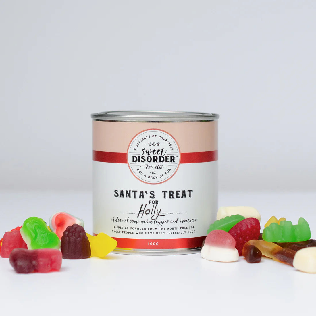 A can with the label "Sweet Disorder" and "Santa's Treat For ... Lollies," surrounded by an assortment of delightful gummy shapes such as bears, rings, and worms. Adorned with red and white design elements, this North Pole formula is crafted to inspire extraordinary moments. Personalize your gift today!