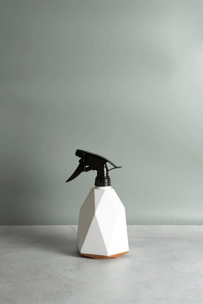 A white geometric spray bottle with a black nozzle, branded as "Ready, Set, Grow" by giftbox co., sits on a light gray surface against a soft gray-green background. The bottle is specifically designed for indoor plant food and features a faceted design that lends it a modern, angular appearance. The lighting casts subtle shadows, enhancing its minimalist aesthetic.