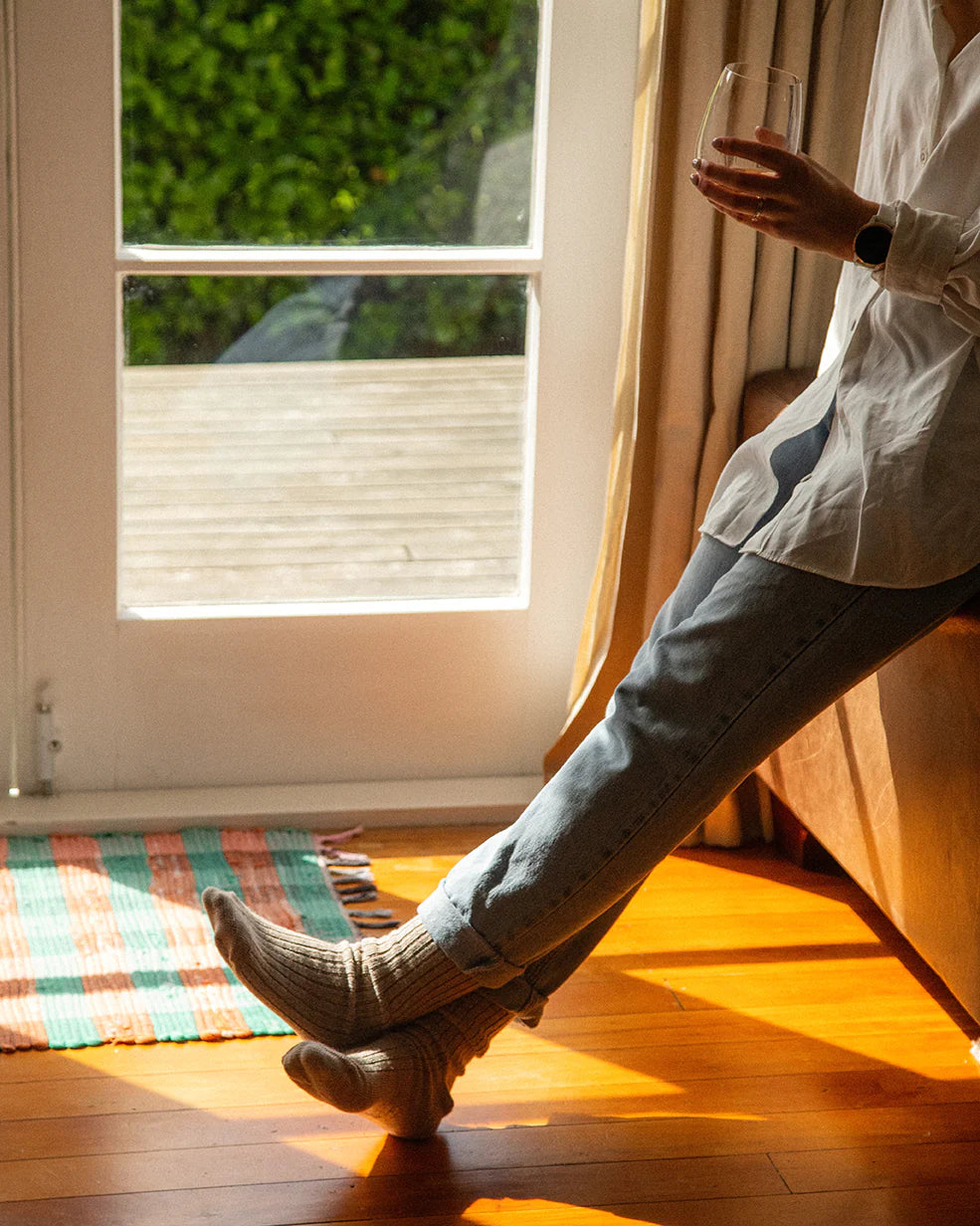 A person wearing a white shirt and light blue jeans sits by a sunlit window, holding a glass with a drink. Their feet, clad in Pretty Fly's Merino Wool Socks, rest on a hardwood floor. A colorful striped rug lies nearby, and lush greenery is visible outside the window.