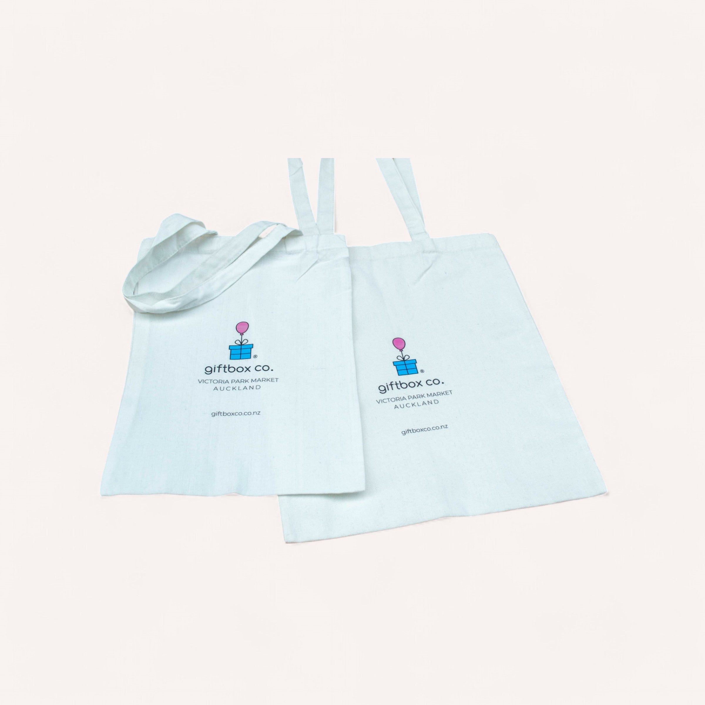 On a plain backdrop, two tote bags in white are displayed. Each bag showcases a vibrant logo featuring a gift box and balloon design, accompanied by the text "giftbox co. VICTORIA ARCADE AUCKLAND giftboxco.nz" printed below. The bags come with long handles, and one is partially folded for display.