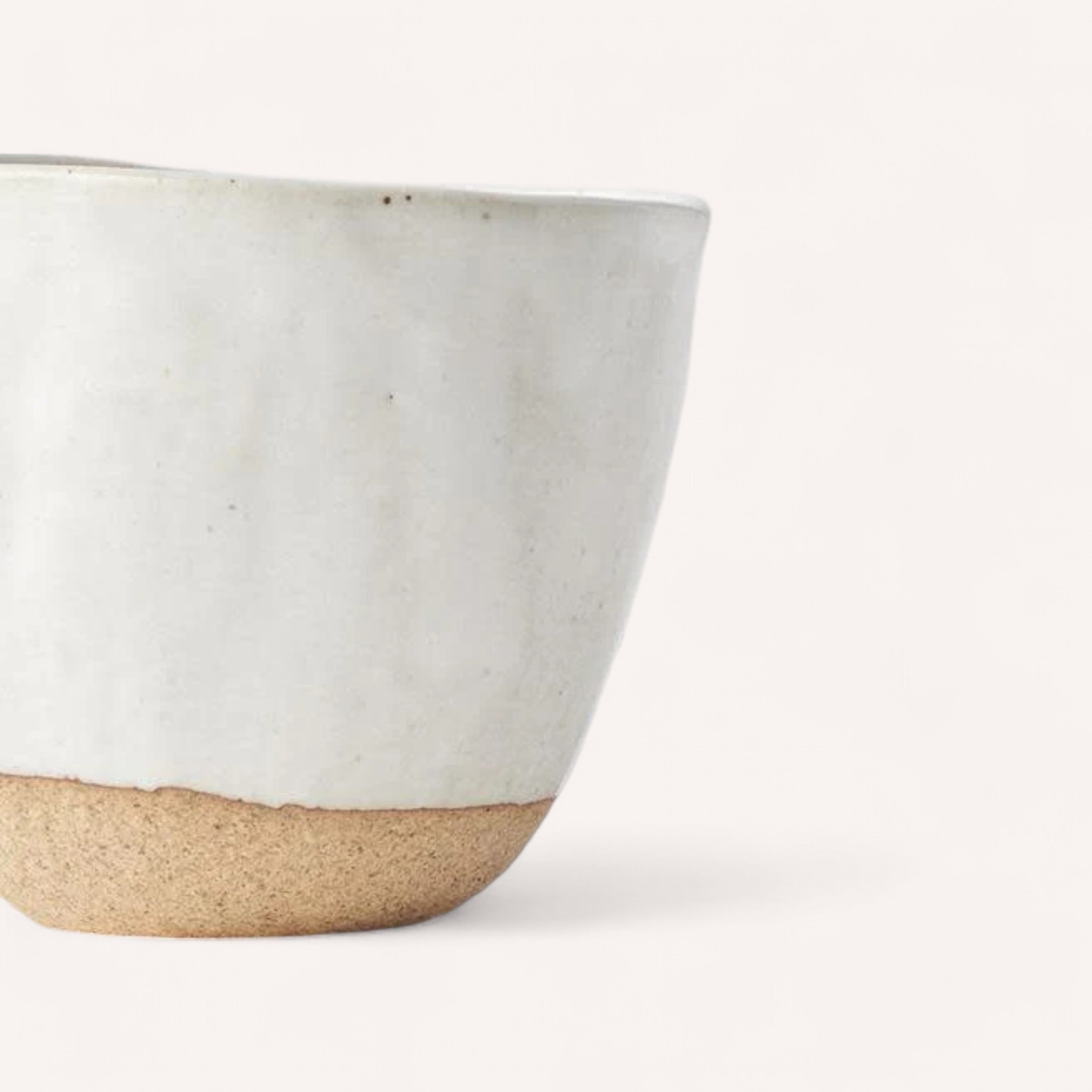 The Naibu Handmade Ceramic Cup showcases a minimalist yet rustic design, featuring a smooth, glossy white glaze over a rough, natural brown clay base. Its hand-finished style includes slight unevenness in shape, complemented by a plain, light background.