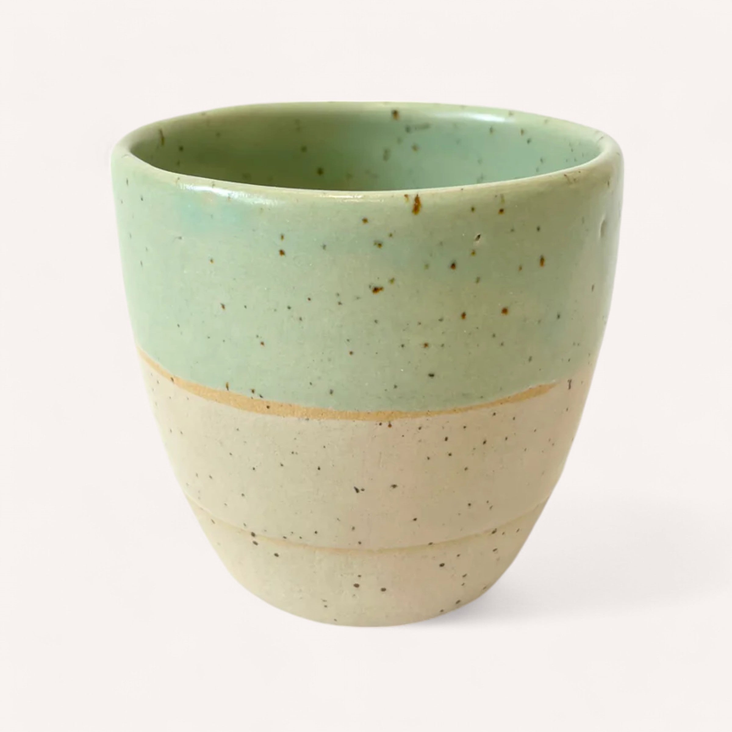 The Handmade Ceramic Cup by Light + Vessel features a glossy mint green upper half and a matte beige lower half, adorned with dark brown speckles. Its smooth, round design is beautifully showcased against a plain white background.