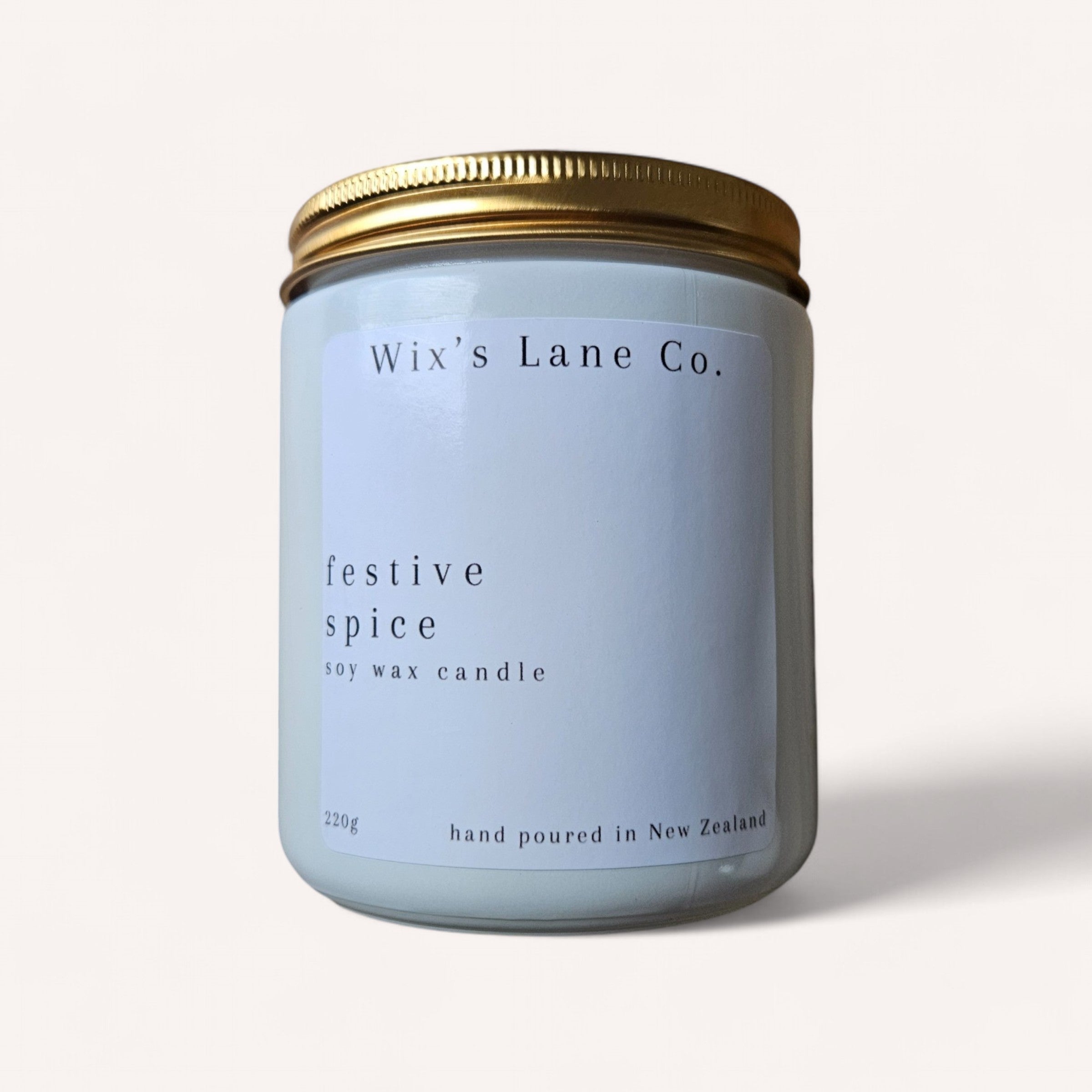 A soy wax candle in a glass jar with a gold metal lid features minimalist design and simple typography on a plain background. The label reads "Wix's Lane Co." and identifies the product as the "Christmas Candle," described as a "festive spice soy wax candle" weighing 220g and hand-poured in New Zealand.